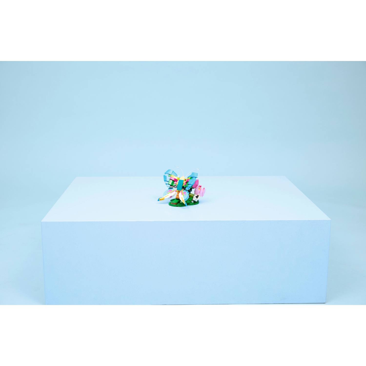 Una scultura di farfalla colorata su un cubo bianco contro uno sfondo azzurro. La farfalla è fatta di blocchi colorati.