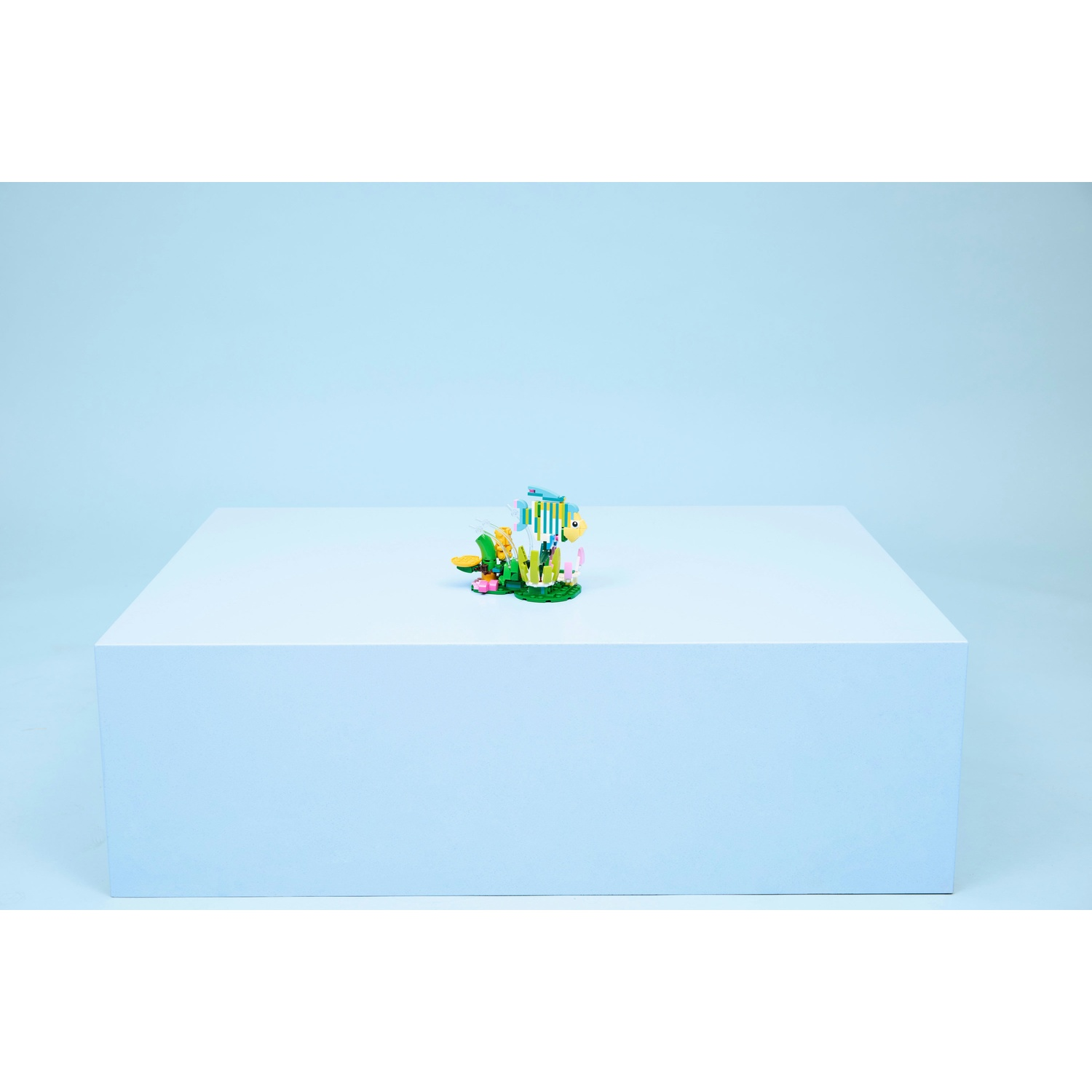 Una piccola scena giocattolo su una piattaforma bianca. Include una recinzione, fiori e un personaggio. Lo sfondo è azzurro.