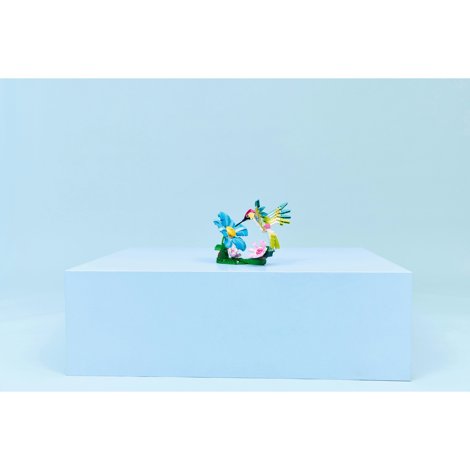 Una statuetta di colibrì colorata su un blocco bianco contro uno sfondo azzurro. È appollaiato su fiori.