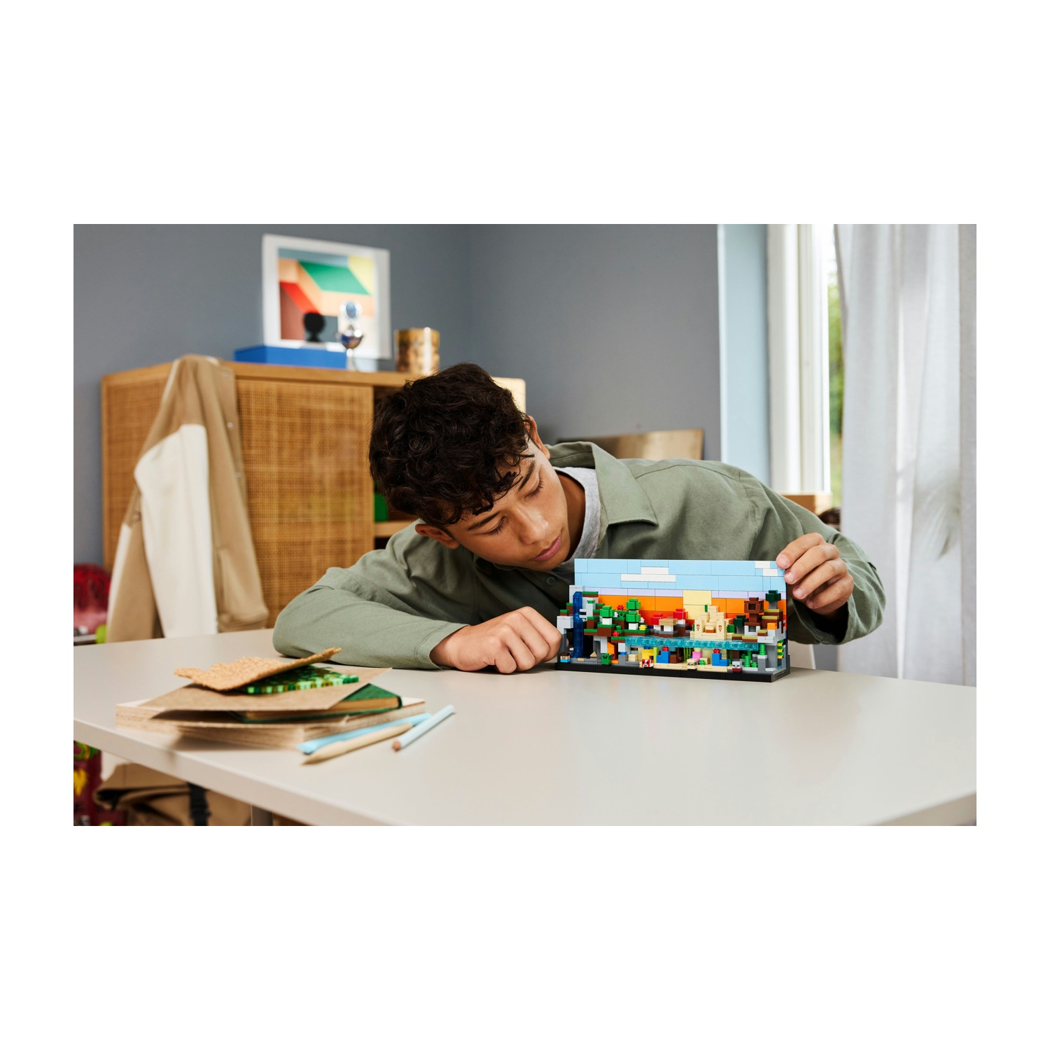 Un giovane si china su un tavolo, guardando un set Lego. Il set raffigura un paesaggio. Altri oggetti, come libri e matite, sono sul tavolo.