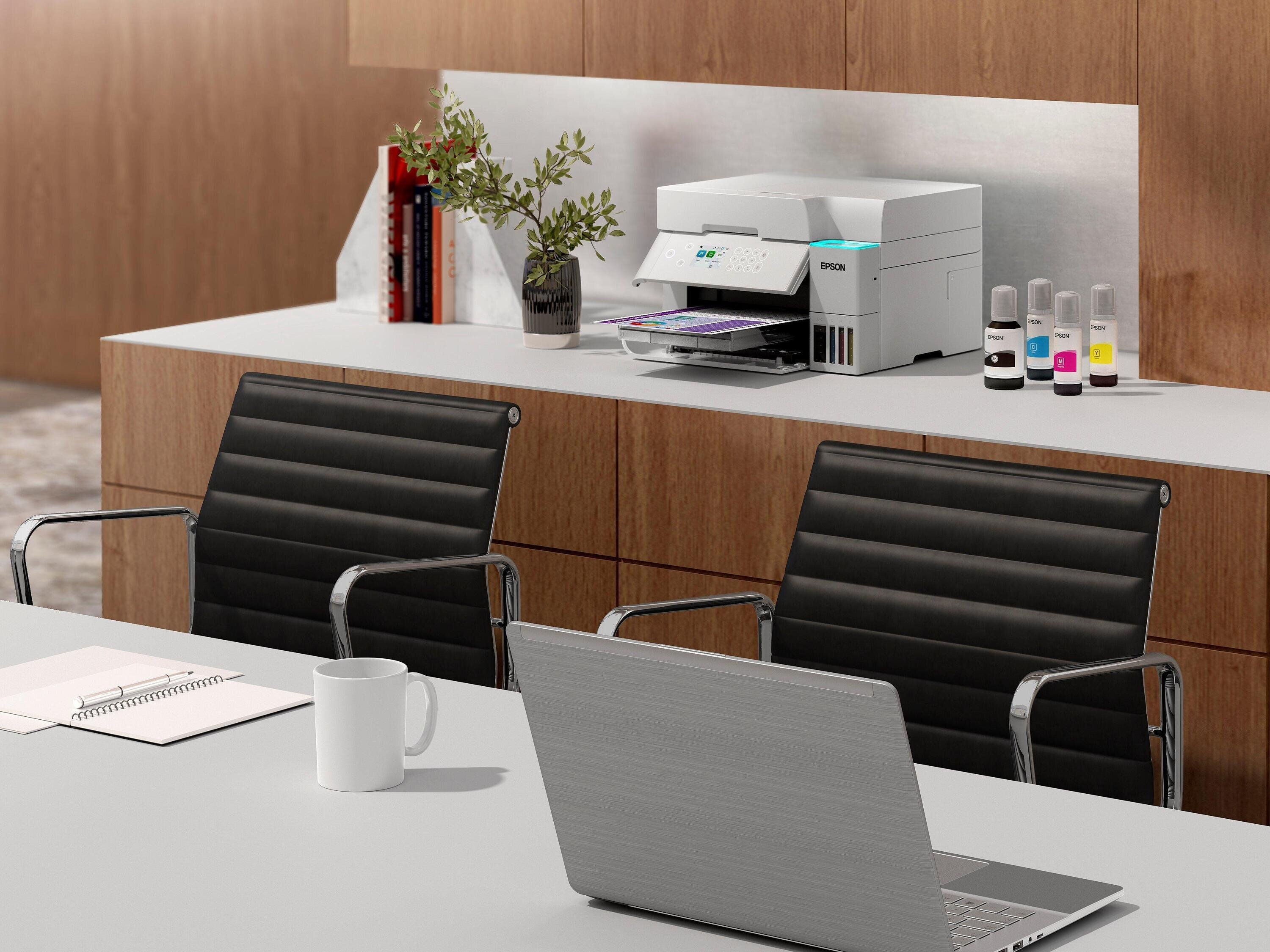 Ustawienie biurowe z drukarką, butelkami z tuszem, laptopem, kubkiem, notatnikiem i dwoma czarnymi krzesłami.