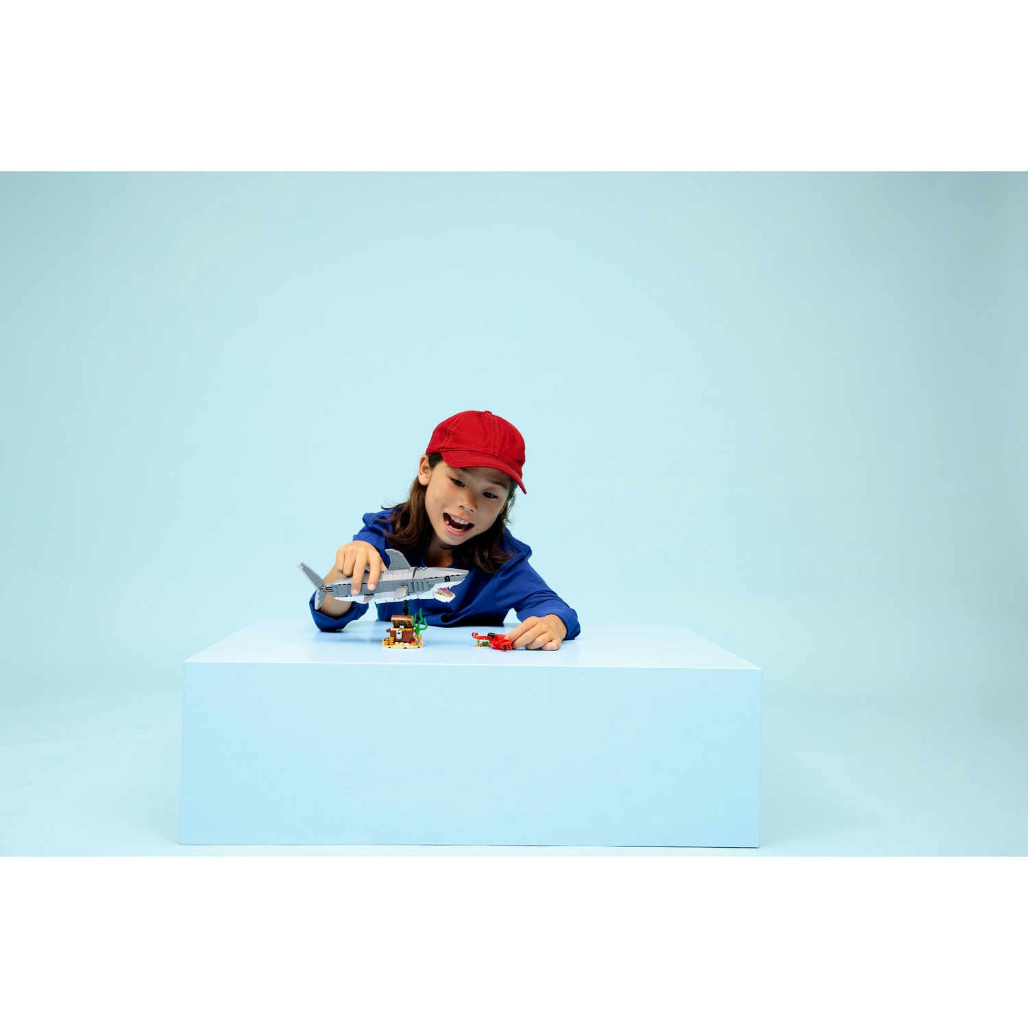 Un bambino con un cappello rosso gioca con giocattoli, tra cui uno squalo, su una piattaforma blu contro uno sfondo azzurro.
