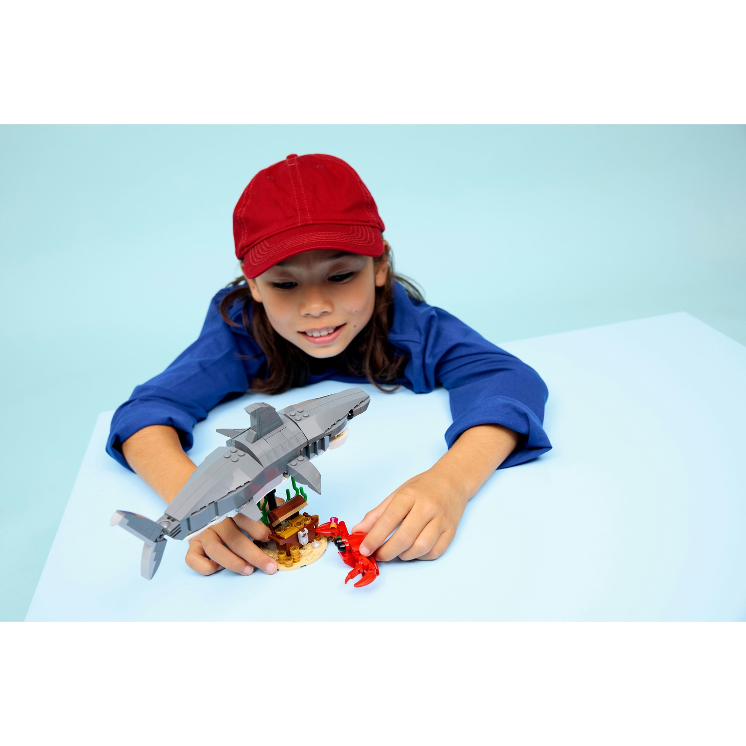 Un bambino con berretto rosso e camicia blu gioca con i Lego: uno squalo, una piccola isola e un granchio su un tavolo blu, sfondo azzurro.