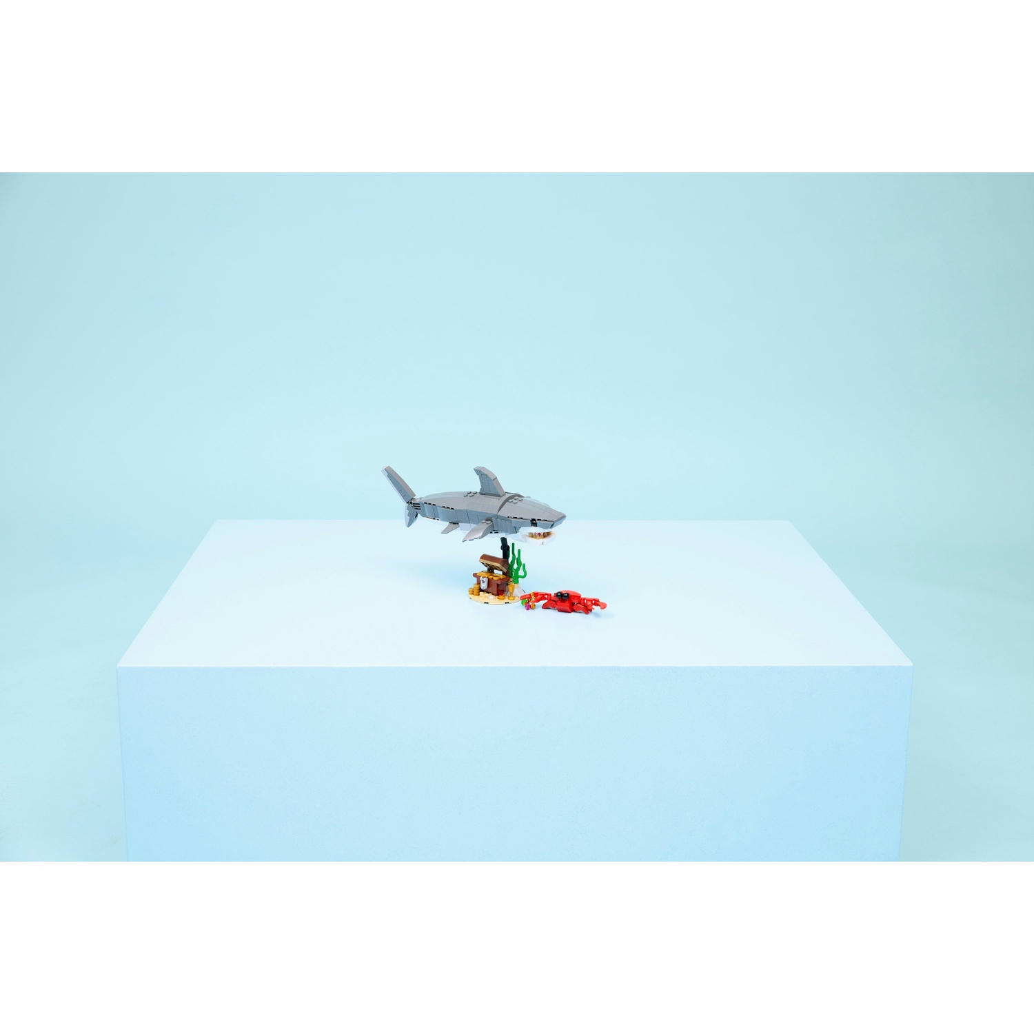 Uno squalo Lego sopra una piccola scena con uno scrigno e un granchio rosso su un blocco bianco su sfondo azzurro.