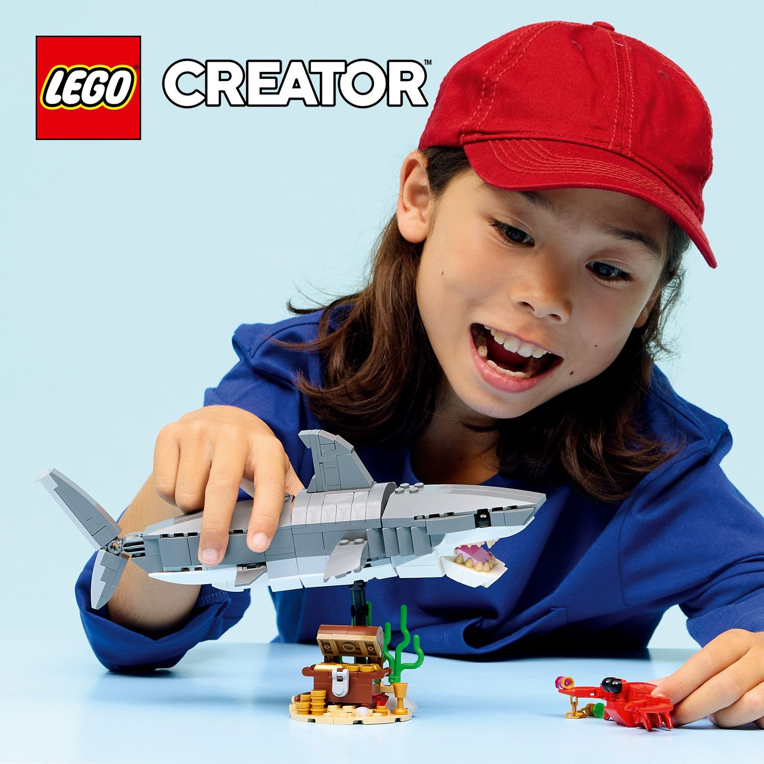 Un bambino con un berretto rosso gioca con lo squalo LEGO Creator, un tesoro e un granchio rosso. Lo sfondo è azzurro.