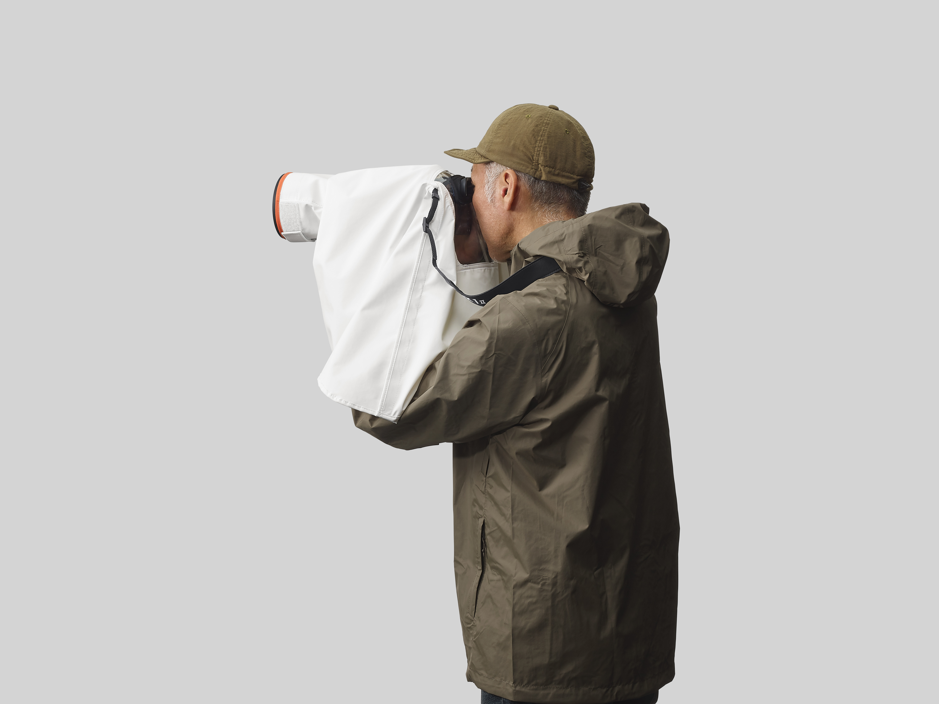 Un uomo con giacca e berretto verdi usa una fotocamera con una copertura bianca. Sfondo grigio.