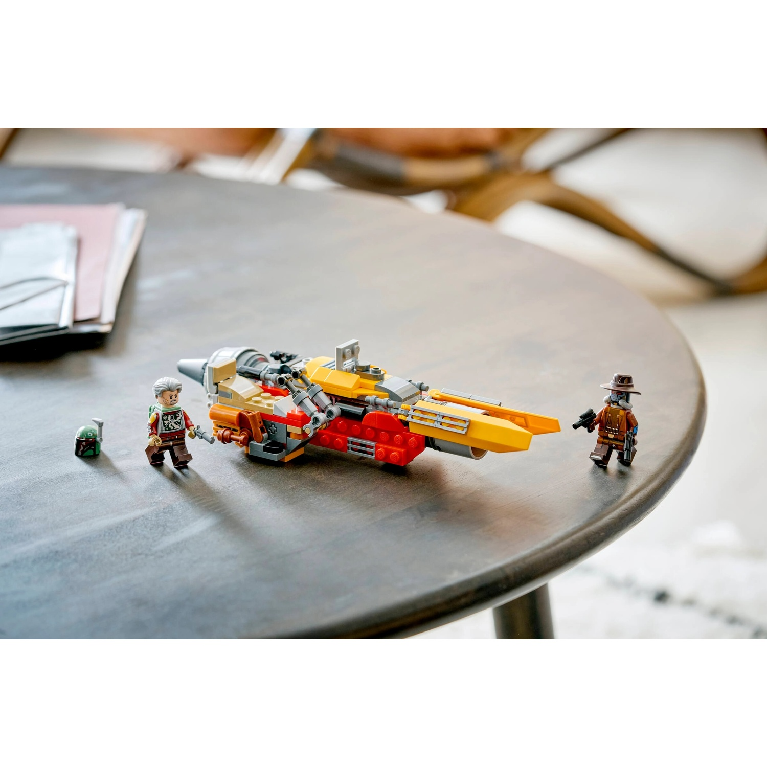 Set Lego su un tavolo. Include una nave gialla e rossa e tre personaggi Lego. Un tavolo grigio e un tappeto bianco sono sullo sfondo.