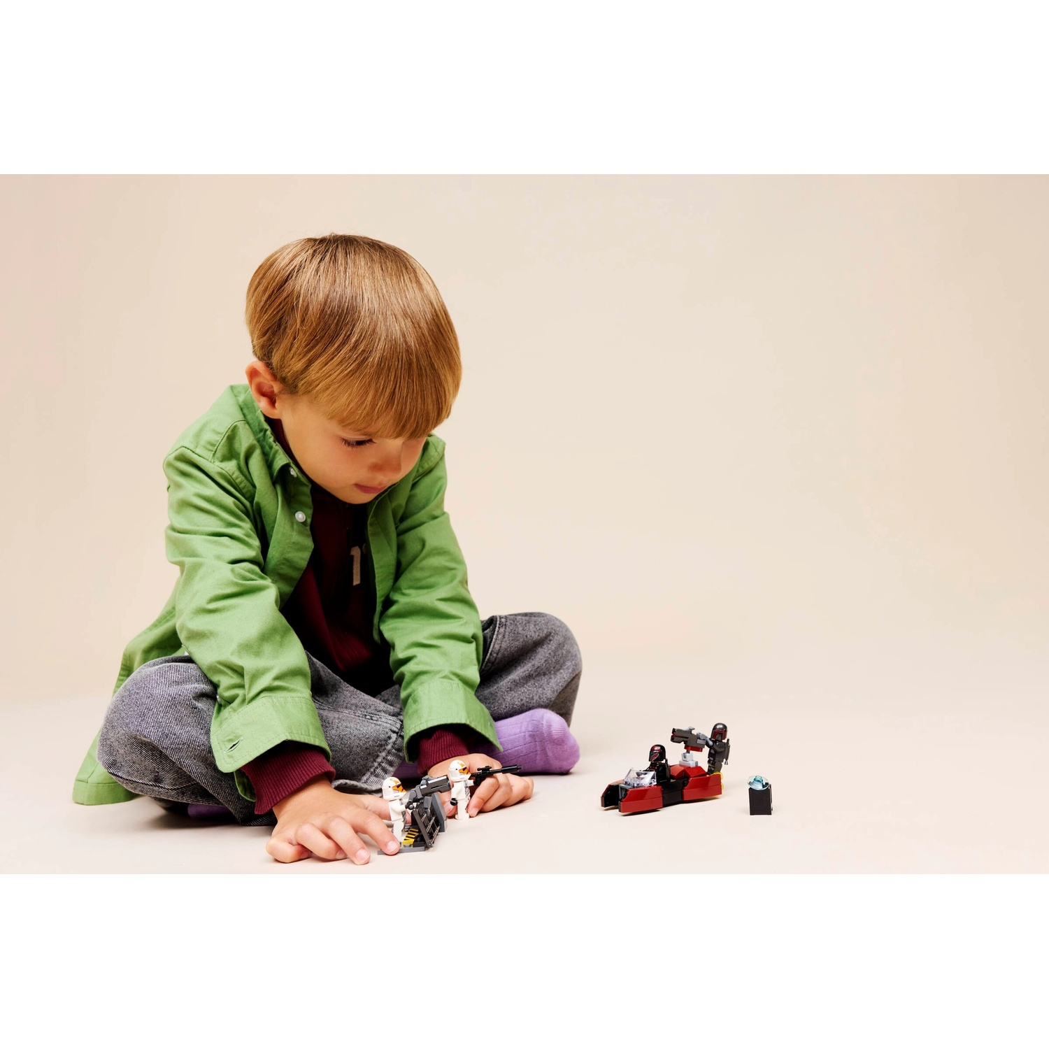 Un bambino seduto gioca con personaggi Lego e un veicolo rosso e nero. Il bambino indossa una camicia verde e pantaloni grigi, calzini viola.
