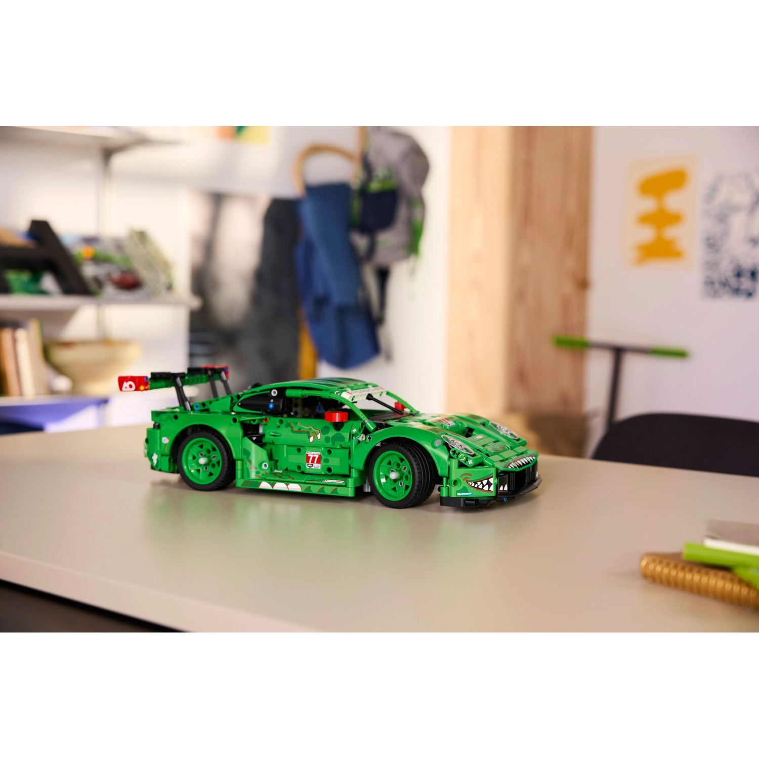 Un'auto da corsa Lego verde è su un tavolo. L'auto ha ruote nere e uno spoiler. Lo sfondo mostra una stanza con una mensola e arte murale.