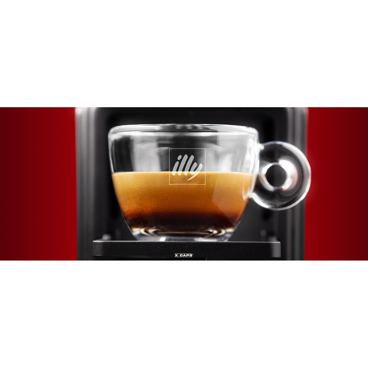 Una tazza di vetro trasparente di caffè espresso con il logo Illy su una superficie nera. Sfondo rosso visibile.