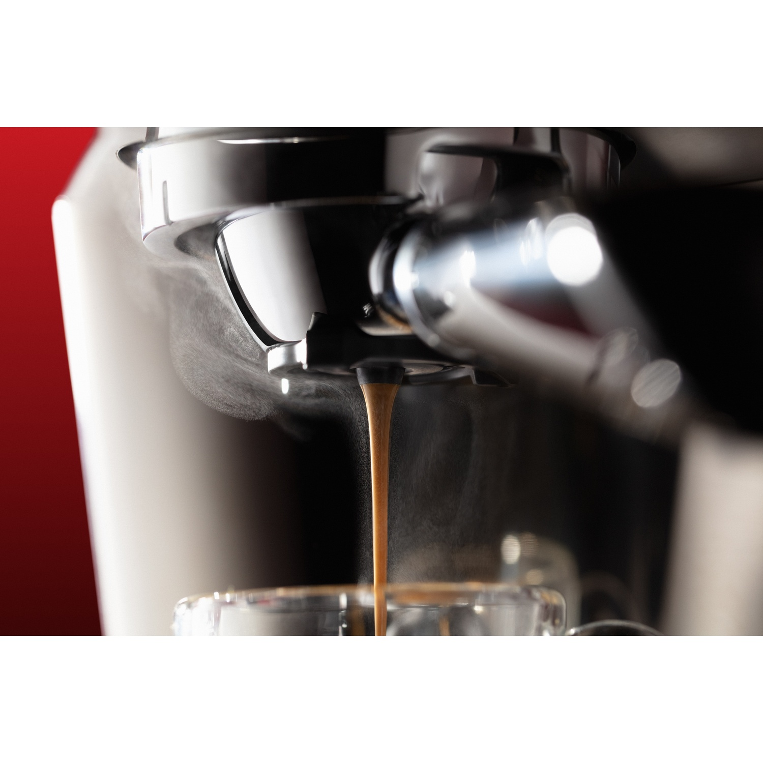 Macchina per caffè espresso che eroga caffè in una tazza di vetro. Il vapore sale. La macchina è in acciaio inossidabile e nera con sfondo rosso.