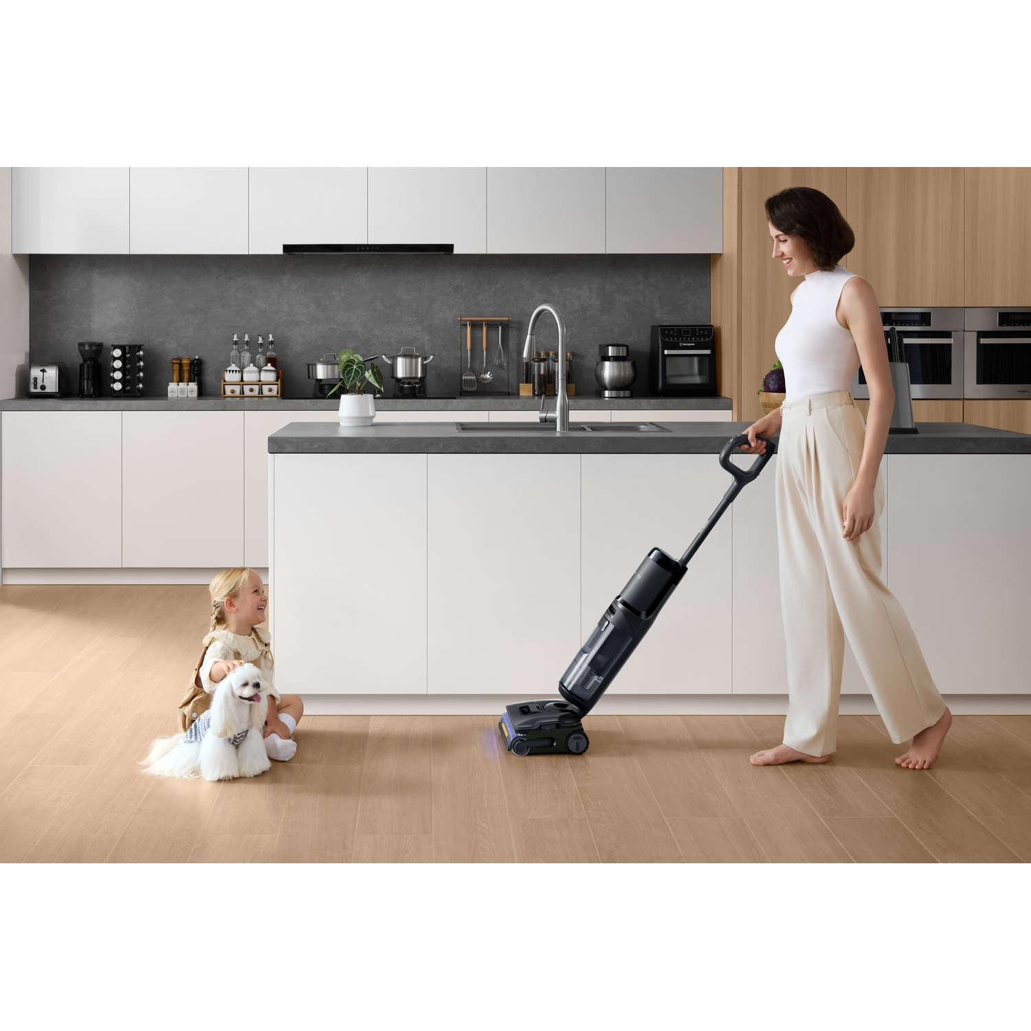 Una donna aspira il pavimento della cucina. Una bambina è seduta con un cane, che guarda. La cucina ha mobili ed elettrodomestici bianchi.