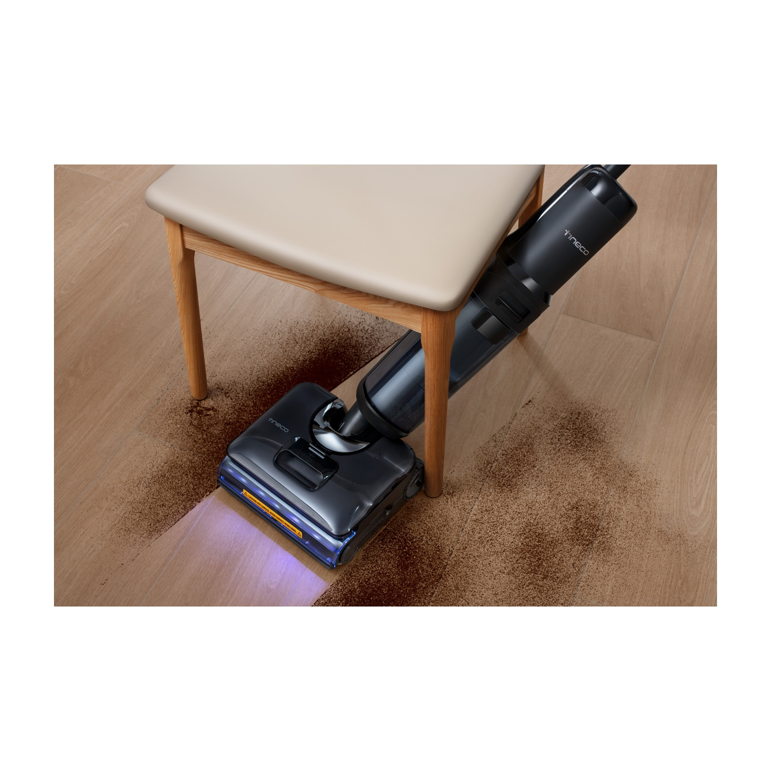 Un aspirapolvere nero pulisce un pavimento in legno coperto di detriti marroni vicino a una sedia con seduta beige.
