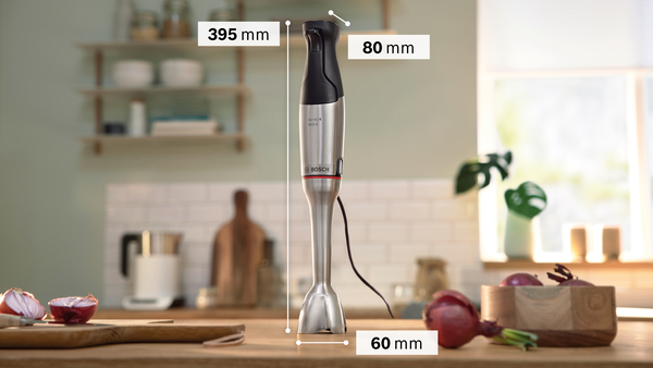 Bir Bosch el blenderi ahşap bir masanın üzerinde duruyor. Ön planda kırmızı soğanlar, bir kesme tahtası ve ahşap bir kase var. Arka plan bir mutfak.