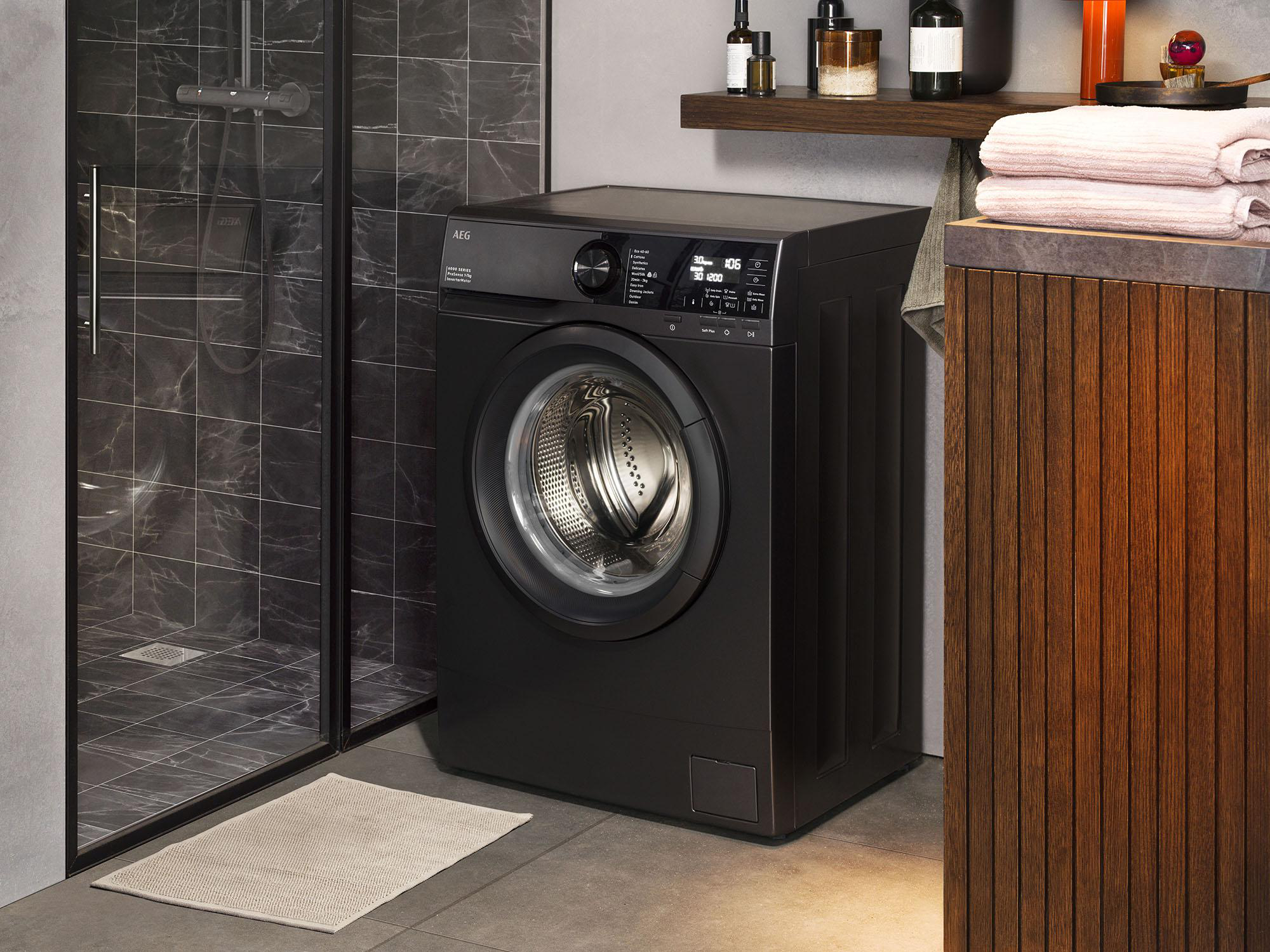 Eine schwarze Waschmaschine in einem Badezimmer. Links eine Duschkabine. Holzschrank mit Handtüchern rechts.