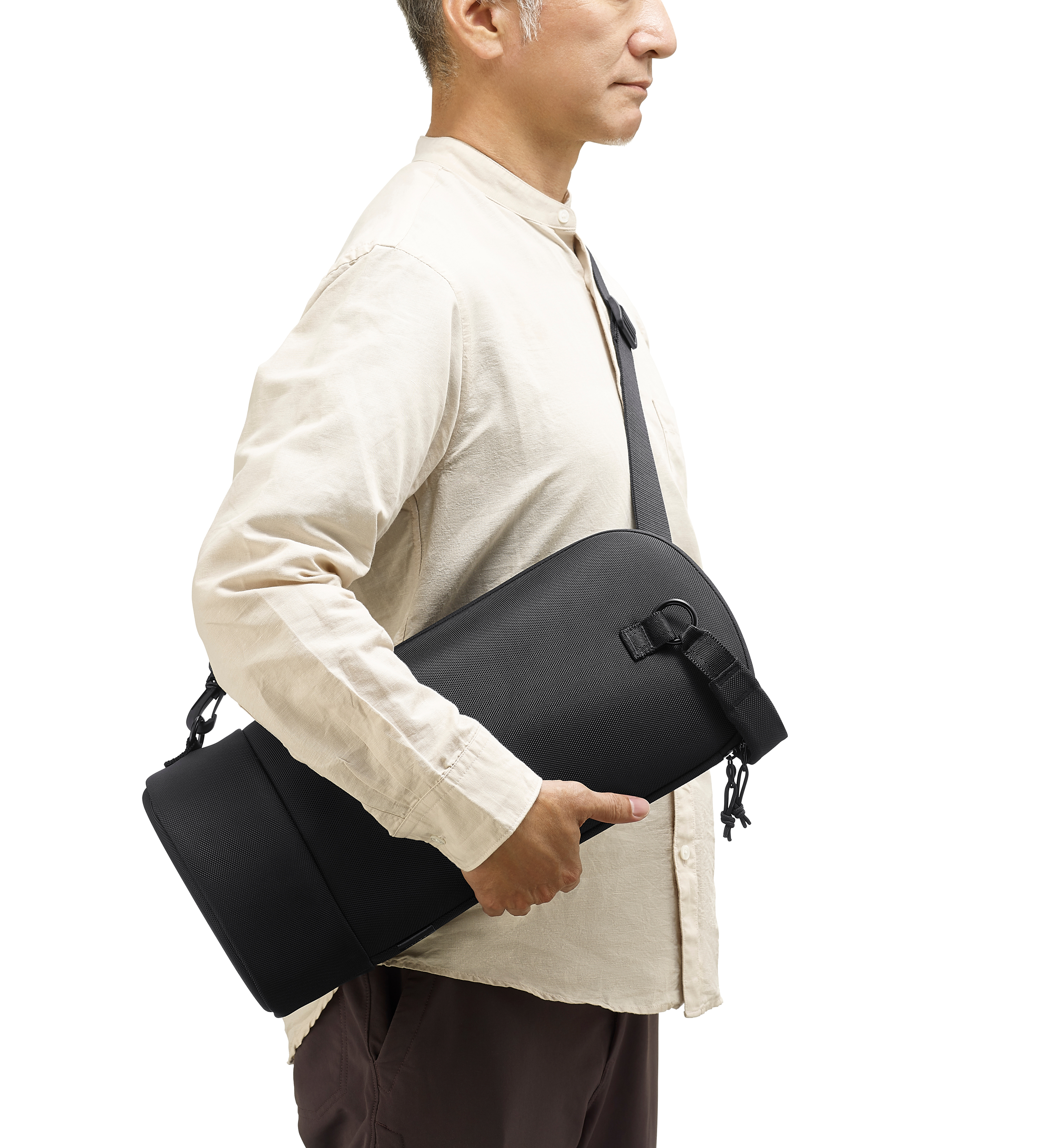 Uomo con borsa nera con tracolla, camicia chiara e pantaloni marroni, su sfondo bianco.