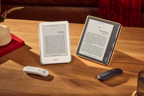 Zwei Kobo E-Reader auf einem Holztisch, die Text anzeigen. Eine weiße und eine schwarze Fernbedienung sind ebenfalls vorhanden, zusammen mit einer Vase.