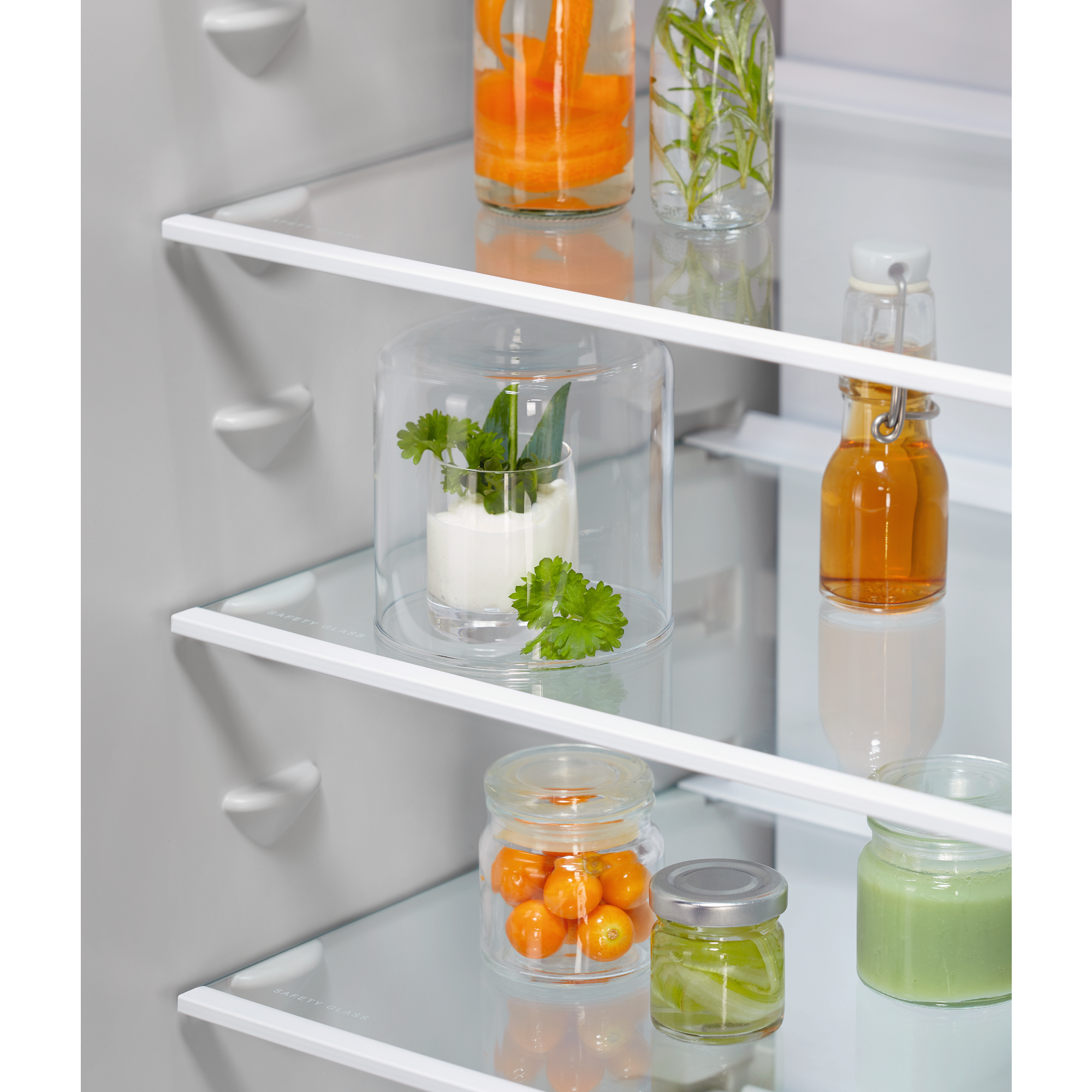 Wewnątrz lodówki widoczne są szklane słoiki z warzywami i ziołami na szklanych półkach. Marchew, zioła i inne składniki.