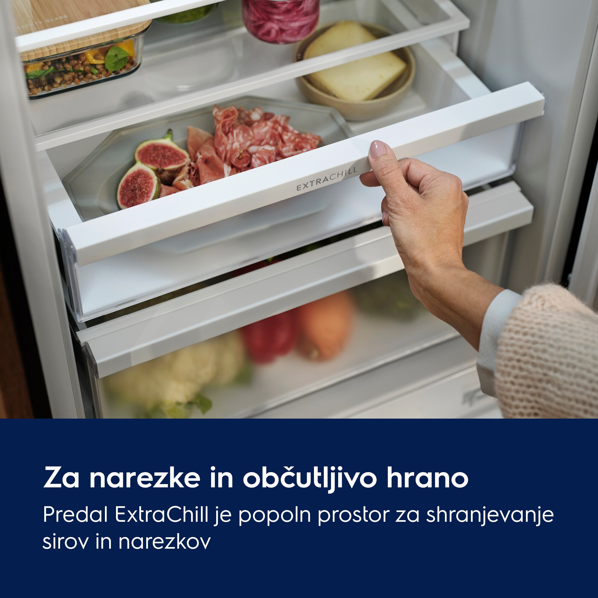 Ręka otwiera szufladę lodówki z napisem ExtraChill, wypełnioną figami i mięsem. Inne szuflady zawierają jedzenie.