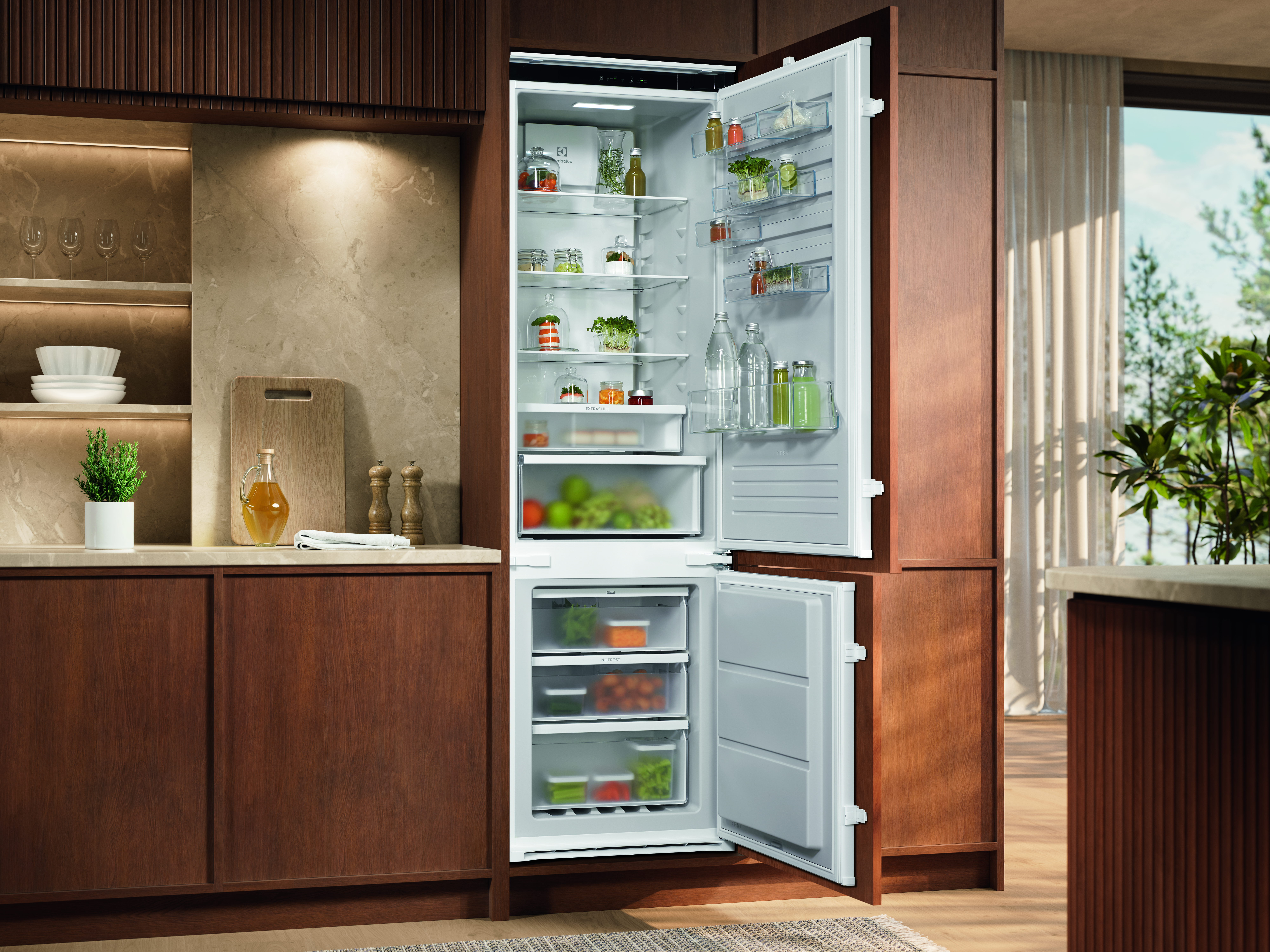 Otwarta lodówka wypełniona jedzeniem. Szafki kuchenne i blat po lewej. Okno i rośliny w tle.