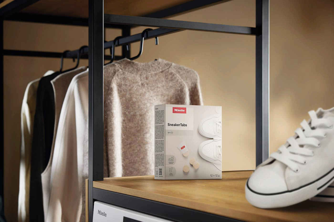 Eine Schachtel Miele SneakerTabs steht auf einem Holzregal neben einem weißen Turnschuh. Kleidung hängt im Hintergrund auf einem Gestell.