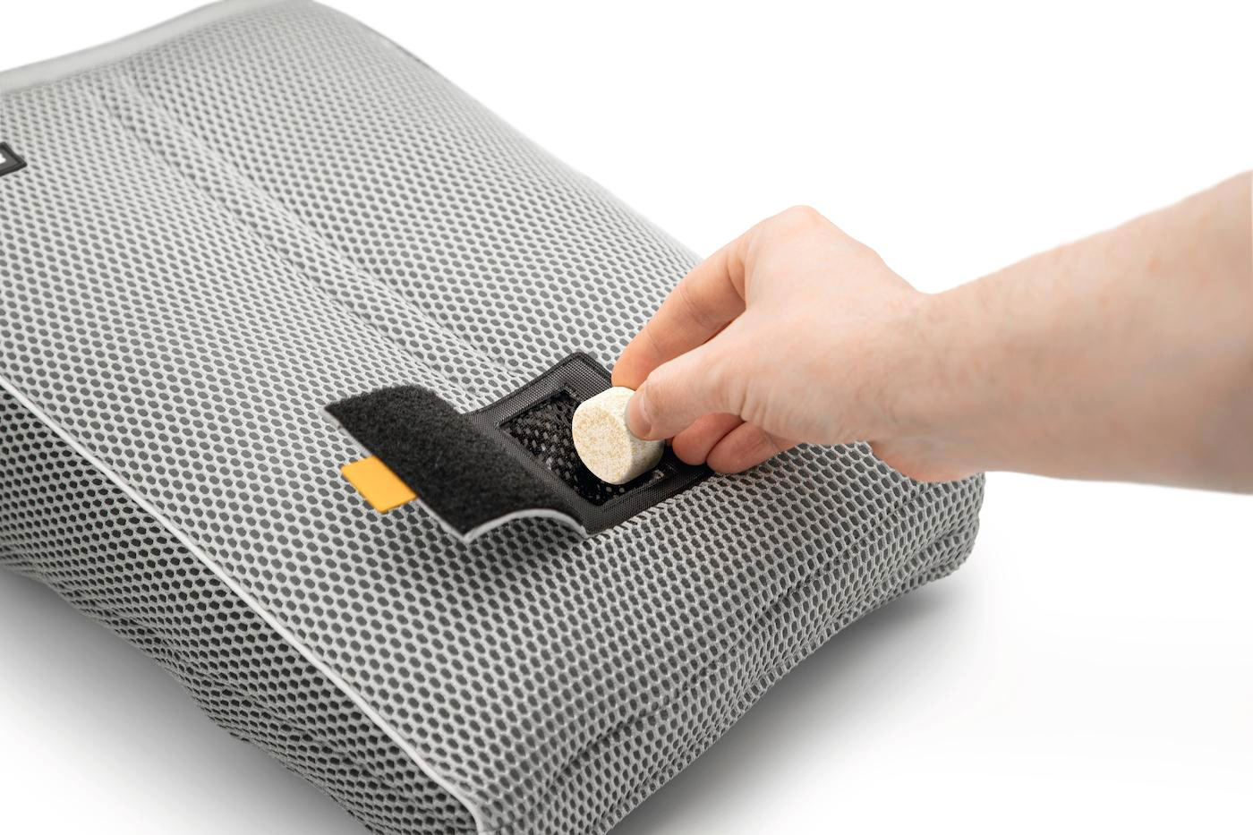 Eine Hand legt einen kleinen, hellbraunen, zylindrischen Gegenstand in eine schwarze Netztasche auf einem grauen Netzpolster. Weißer Hintergrund.
