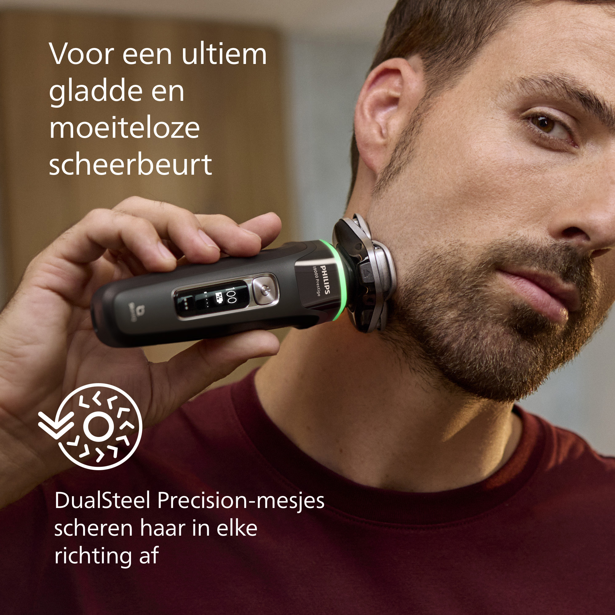 A man shaves his beard with an electric shaver. The shaver is black and green. The man wears a burgundy shirt.