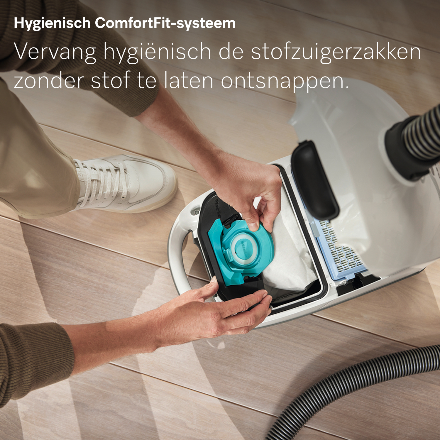 Een persoon vervangt een stofzuigerfilter. De stofzuiger is wit en zwart. Het filter is turquoise en wit, op een houten vloer.