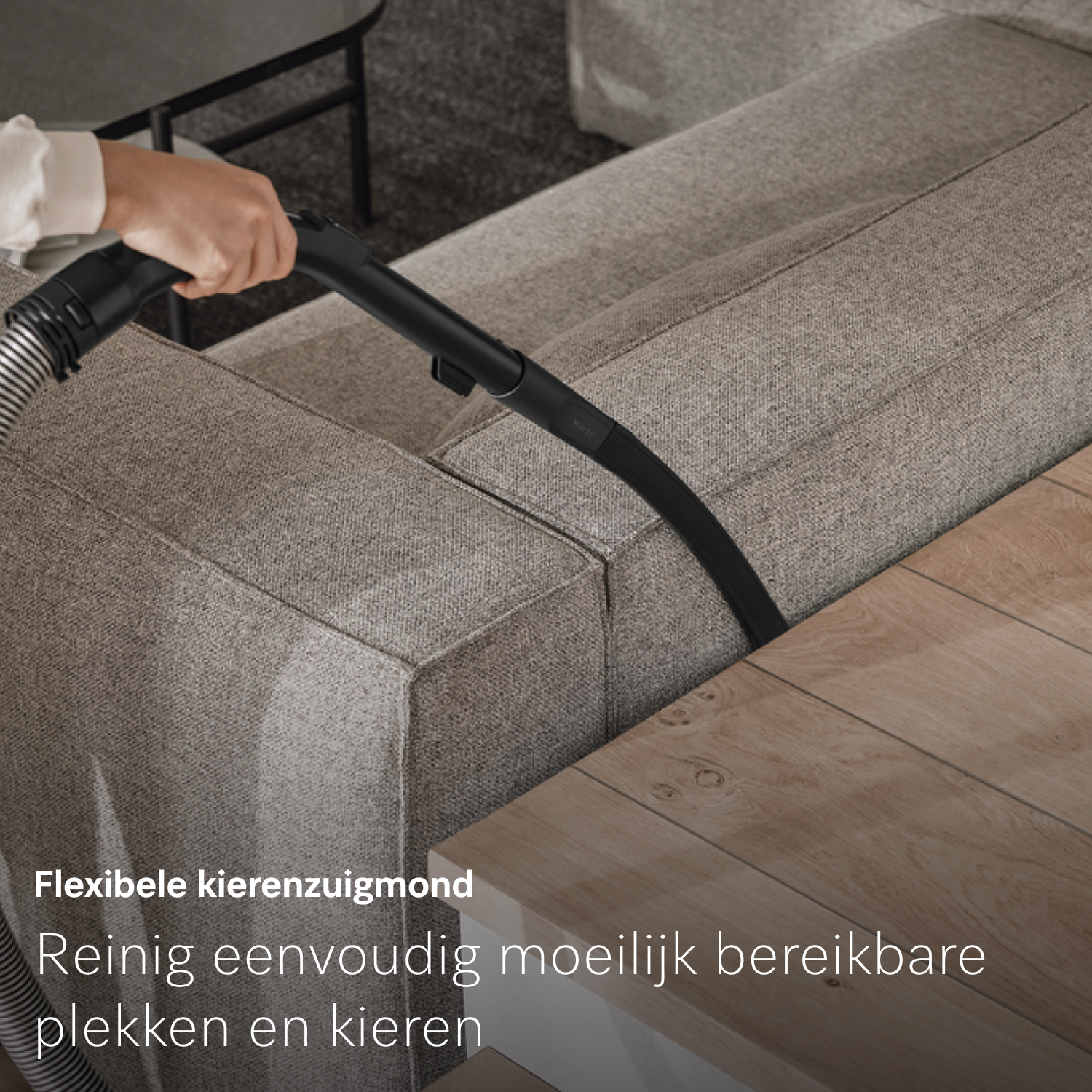Een hand stofzuigt een grijze bank. De slang en het mondstuk van de stofzuiger zijn zwart. Een houten tafel staat op de voorgrond en een bank op de achtergrond.