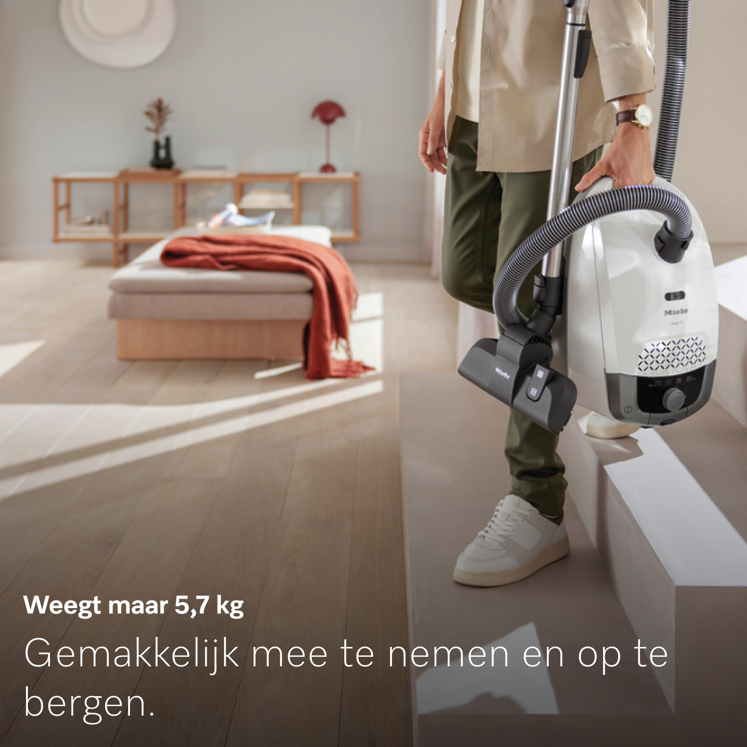 Een persoon houdt een witte Miele stofzuiger vast. Ze staan op een trap. Een bank met een rode deken staat op de achtergrond.
