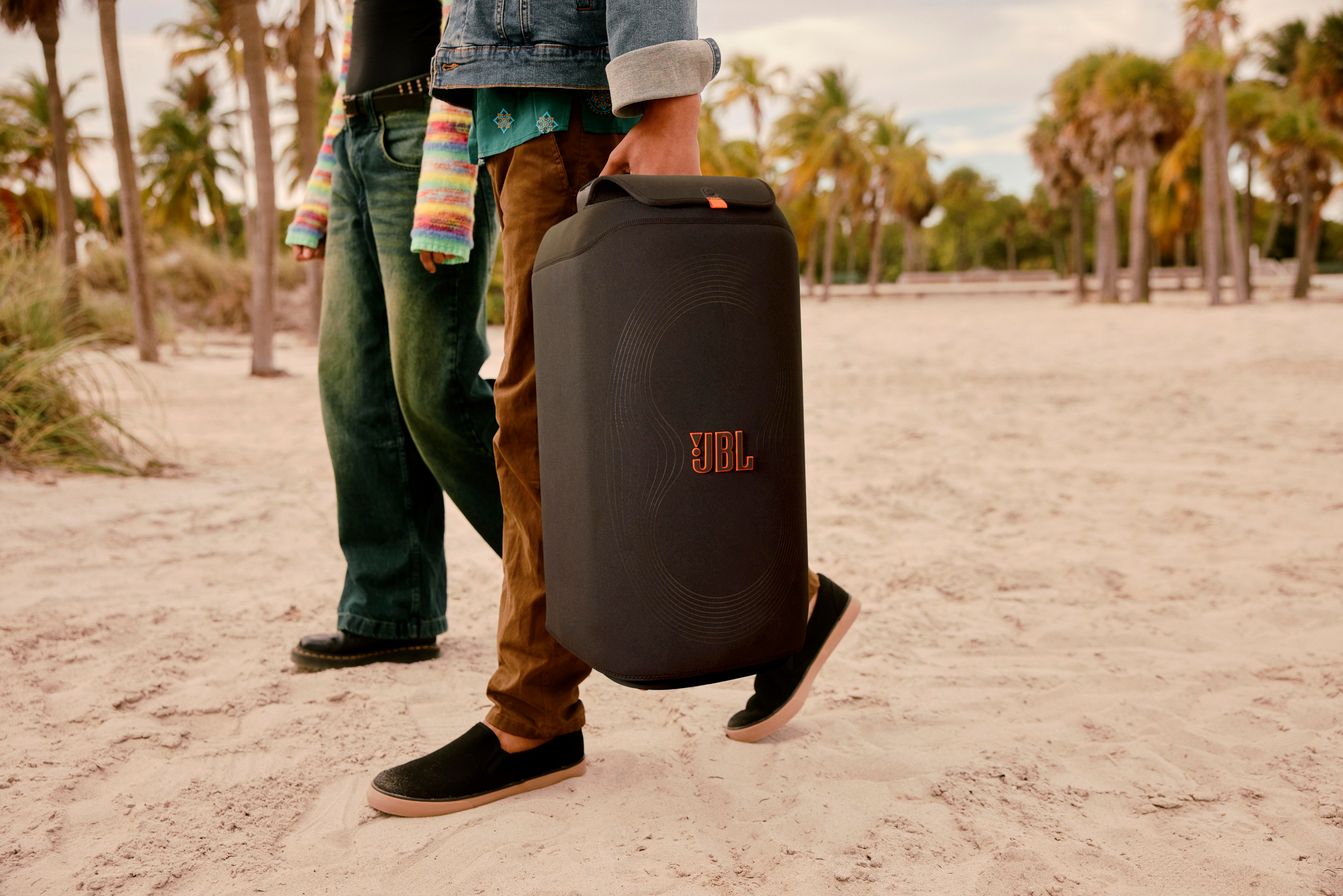 Person am Strand mit schwarzem JBL-Lautsprecher. Palmen im Hintergrund. Zwei weitere Personen in der Nähe.