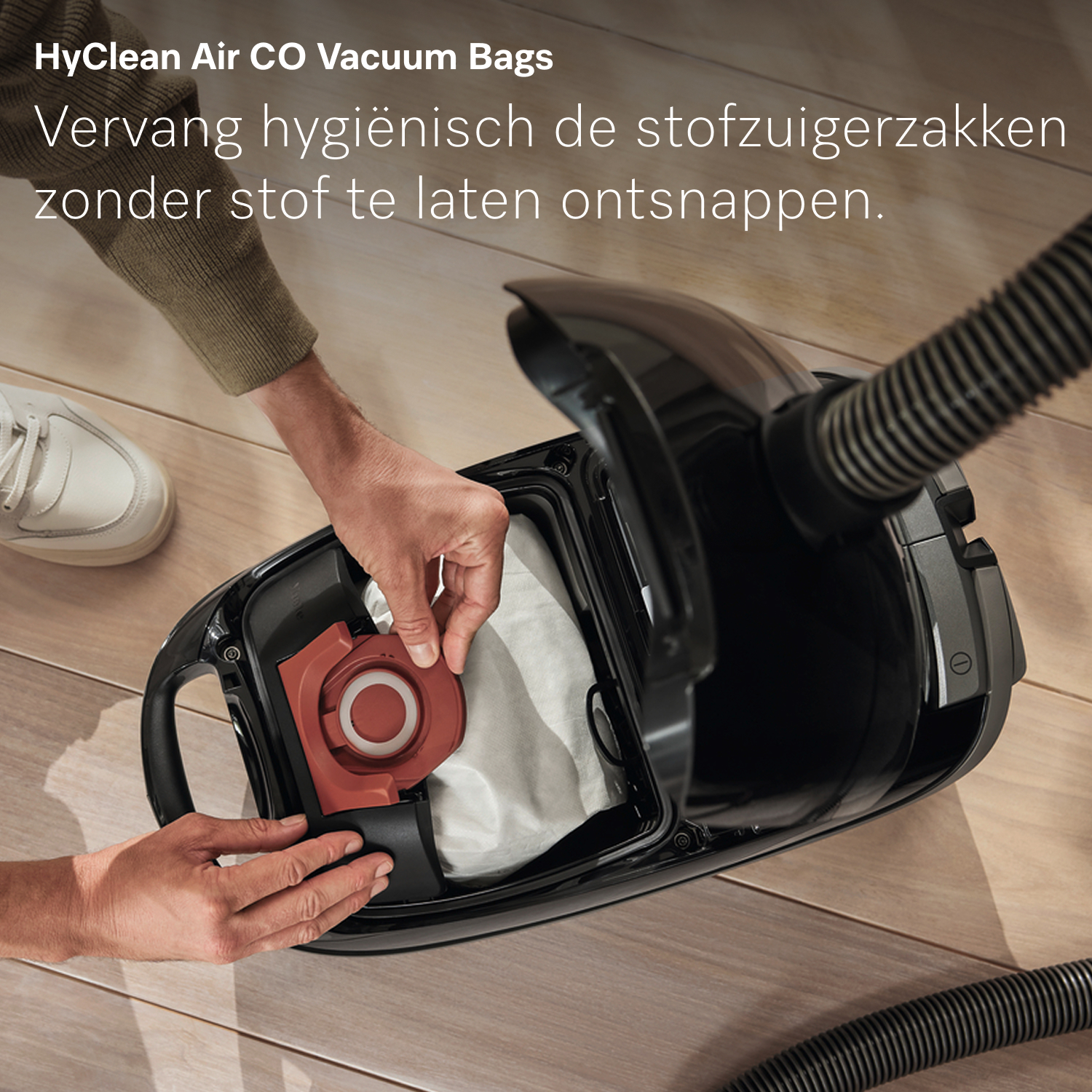 Een persoon vervangt een stofzuigerzak. De stofzuiger is zwart, de zak is wit en de achtergrond is een houten vloer.