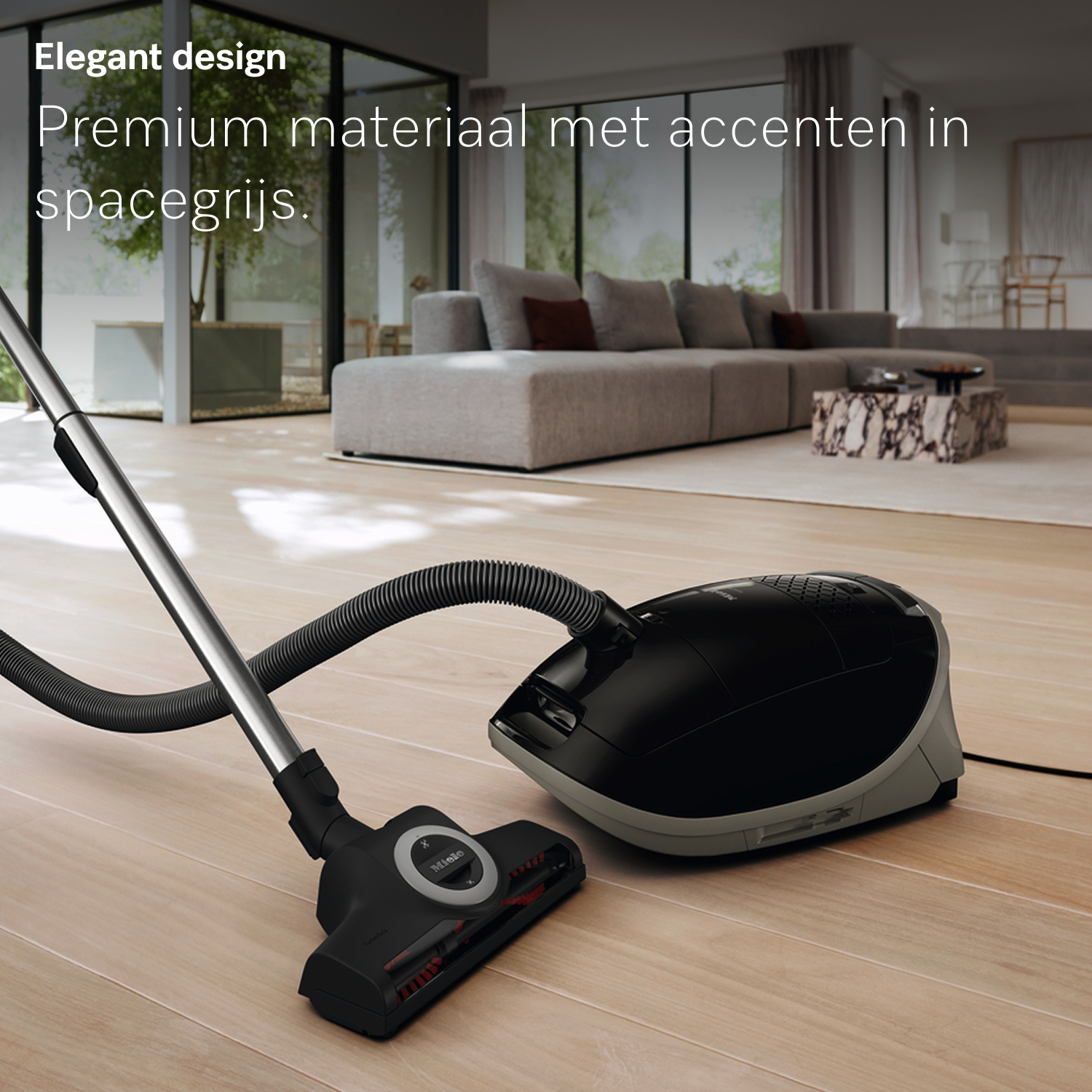A black and gray vacuum cleaner is on a wooden floor. In the background, there is a living room with a gray sofa.