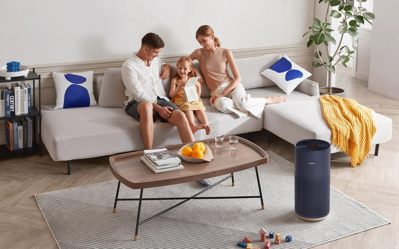 Rodzina na sofie, czytająca książkę. Stół z pomarańczami i szklankami oraz oczyszczacz powietrza w pokoju.