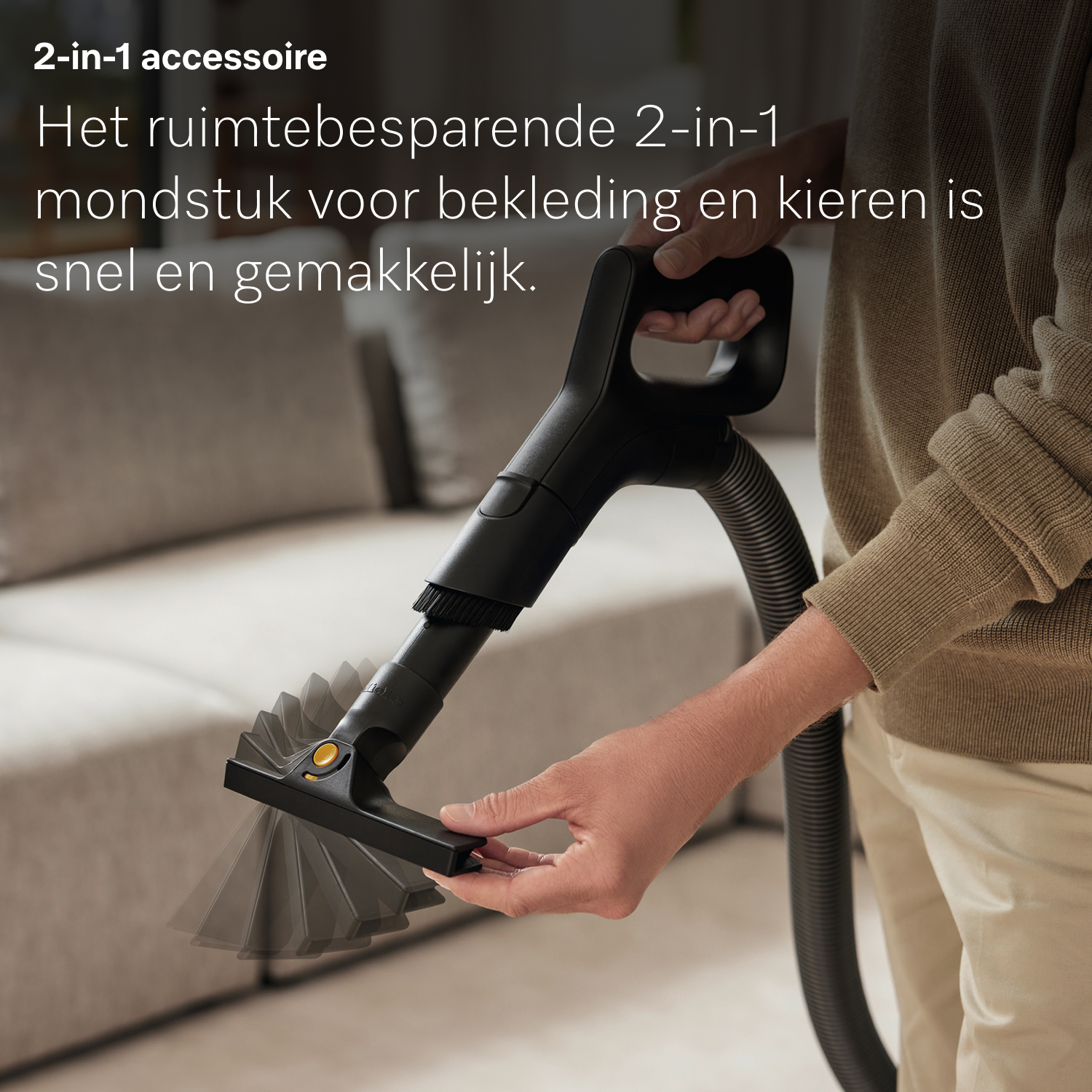 Een persoon houdt een zwart stofzuigeropzetstuk vast. Het opzetstuk is naar de kijker gericht, met een grijze bank op de achtergrond.