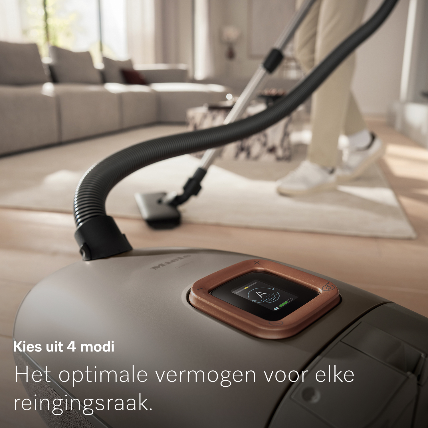 Een persoon stofzuigt een woonkamer met een grijze stofzuiger. De stofzuiger heeft een digitaal display en de woorden 'Kies uit 4 modi' zijn zichtbaar.