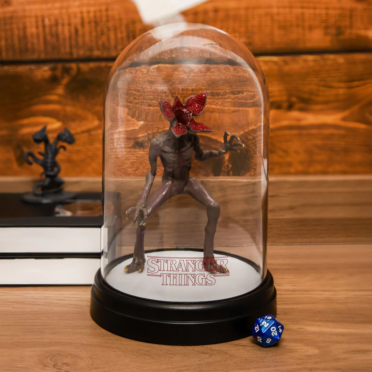 Figura de Demogorgon dentro de una cúpula de cristal, con un dado azul y una pequeña estatua sobre una superficie de madera. El fondo es una pared de madera.