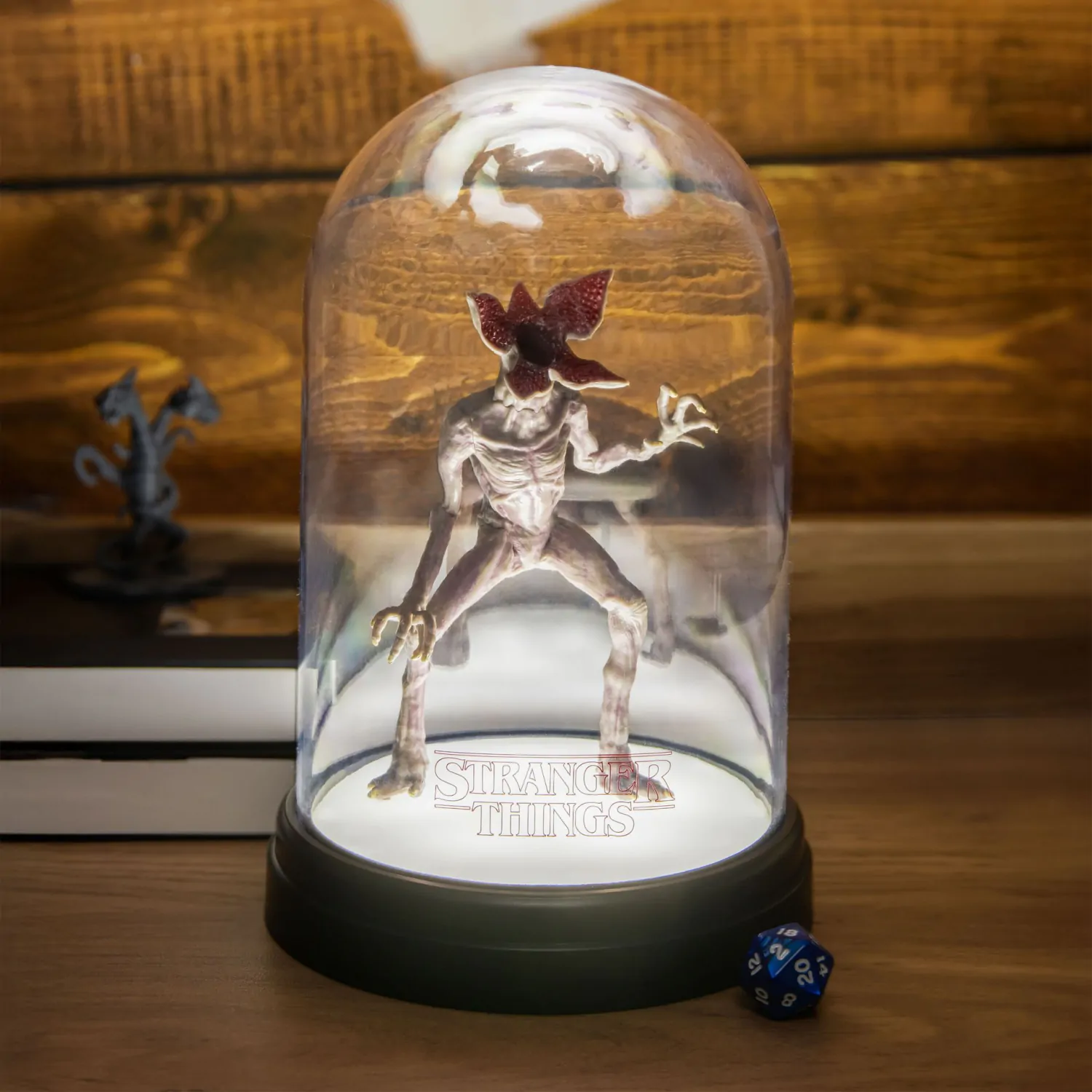 Figura de Demogorgon dentro de una cúpula de vidrio, iluminada con el logotipo de Stranger Things. Fondo de madera con un dado y una pequeña estatua.