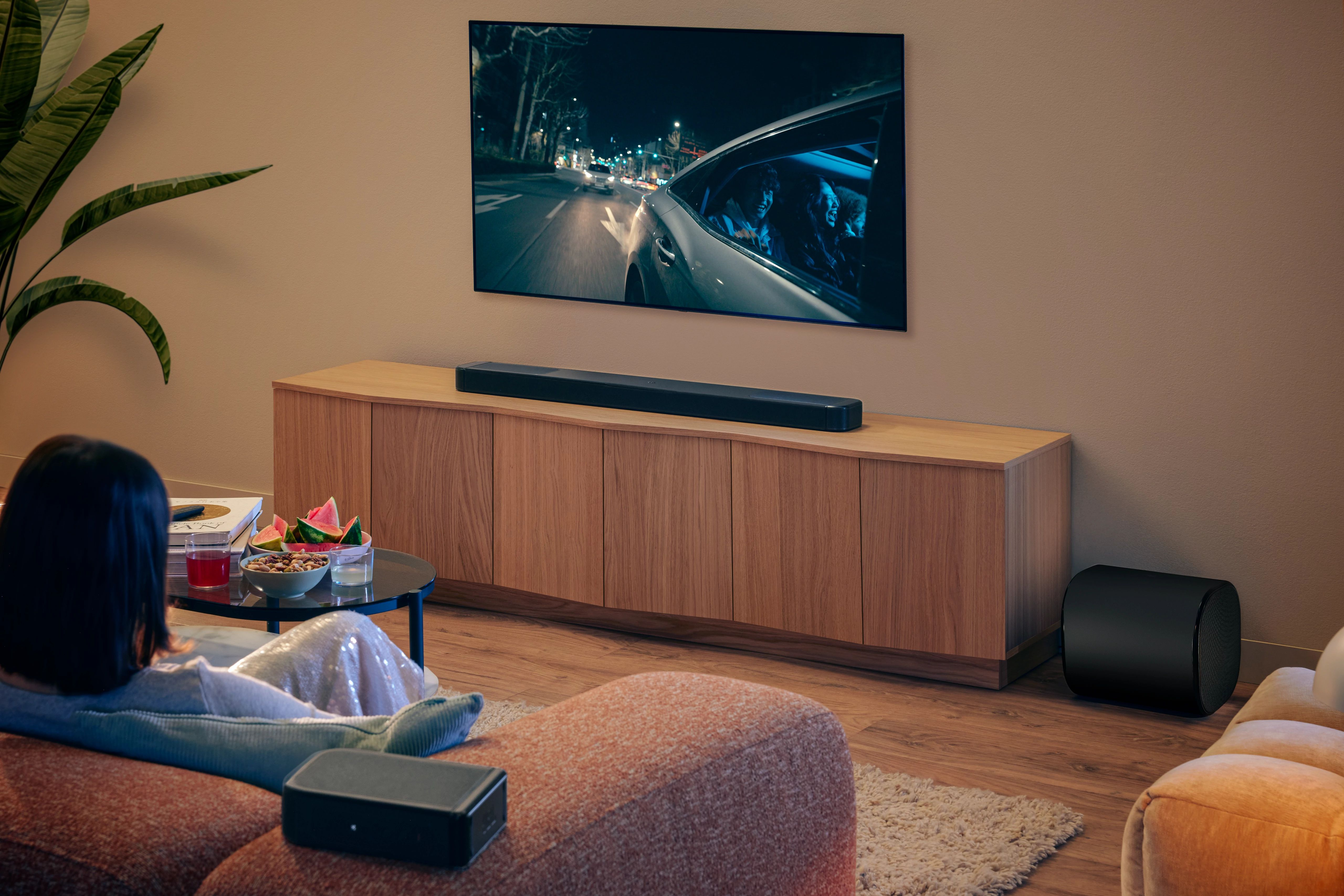 Oturma odası sahnesi. Bir kadın TV izliyor. Soundbar ve subwoofer görülebilir. Masada atıştırmalıklar ve içecekler.