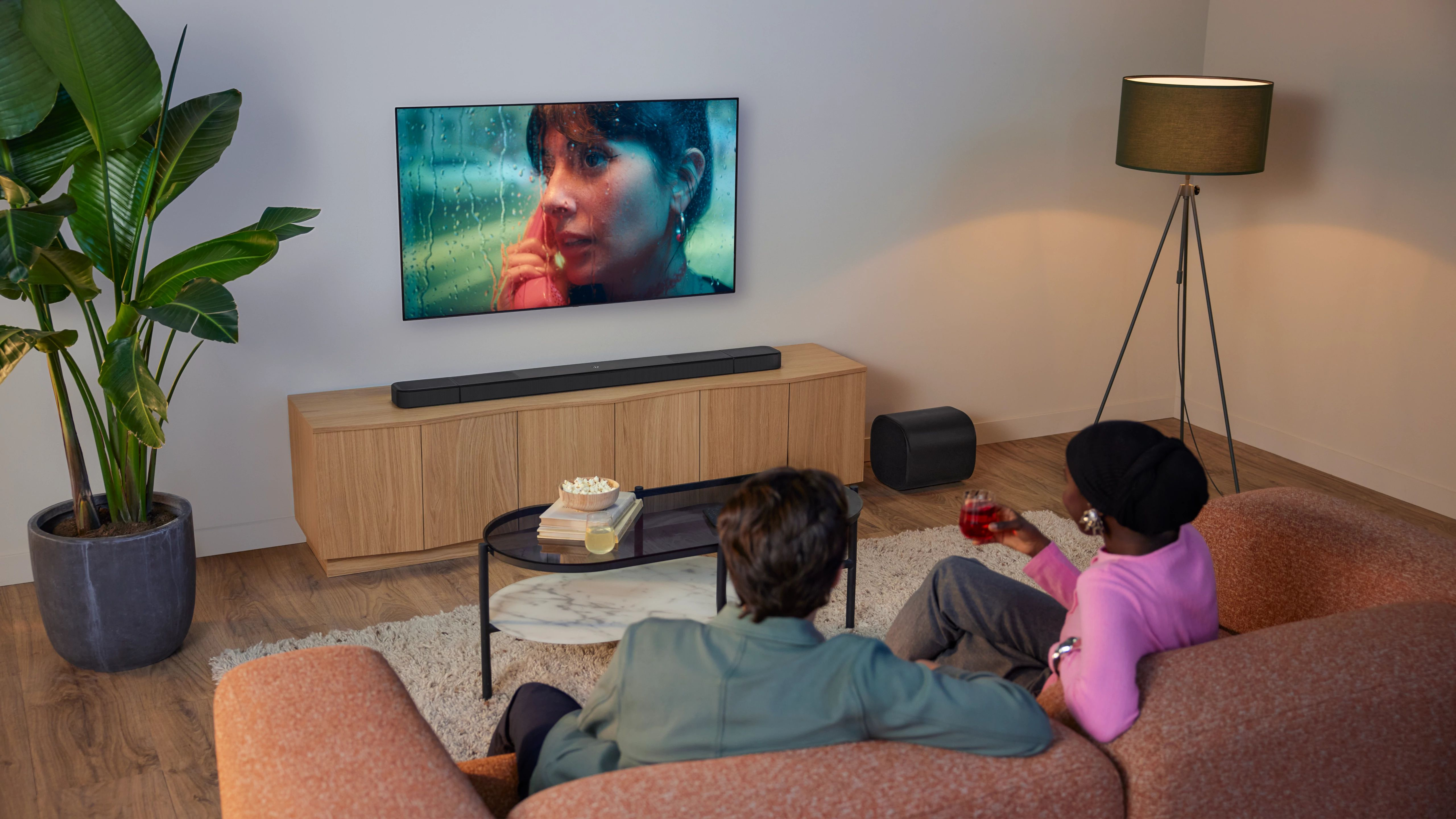 Oturma odası sahnesi. TV'de telefonla konuşan bir kadın var. Kanepe üzerinde bir çift, biri içecek tutuyor. Bitki, lamba ve soundbar görünür.