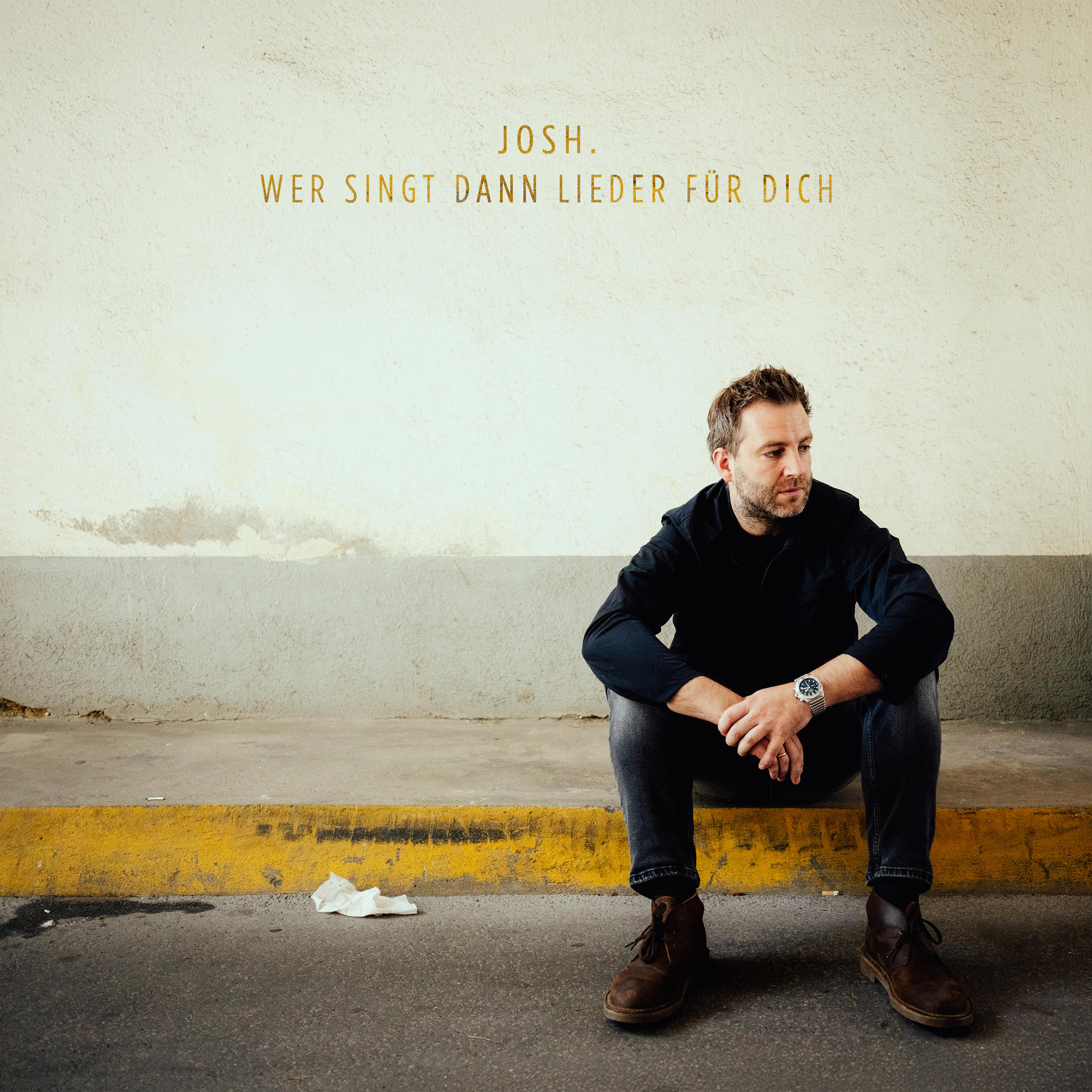 Ein Mann sitzt auf einer gelb gefütterten Leiste und trägt schwarze Kleidung. Der Text oben lautet: JOSH. WER SINGT DANN LIEDER FÜR DICH.