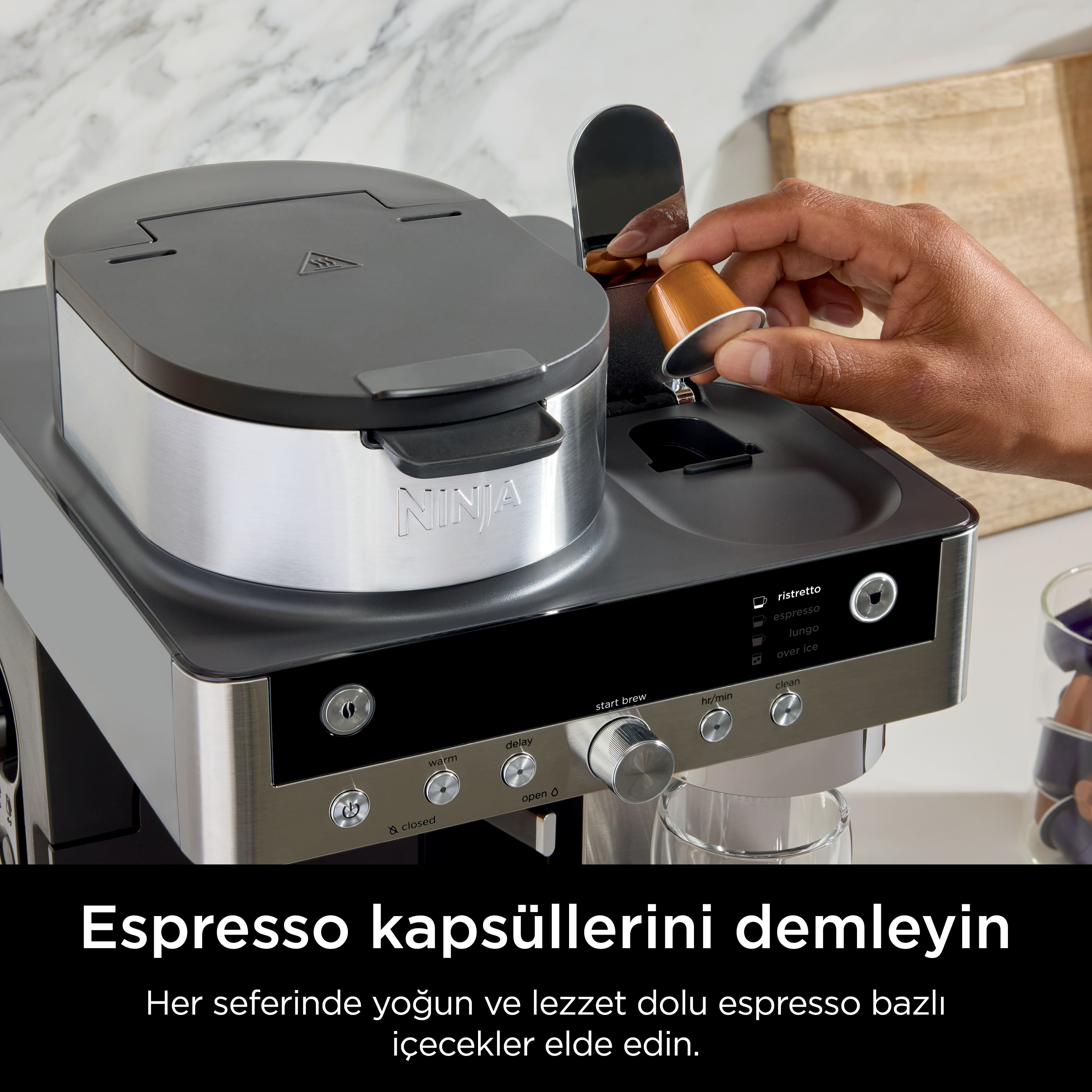 Bir kişi bir Ninja espresso makinesine kapsül yerleştiriyor. Makine gümüş ve siyah renktedir.