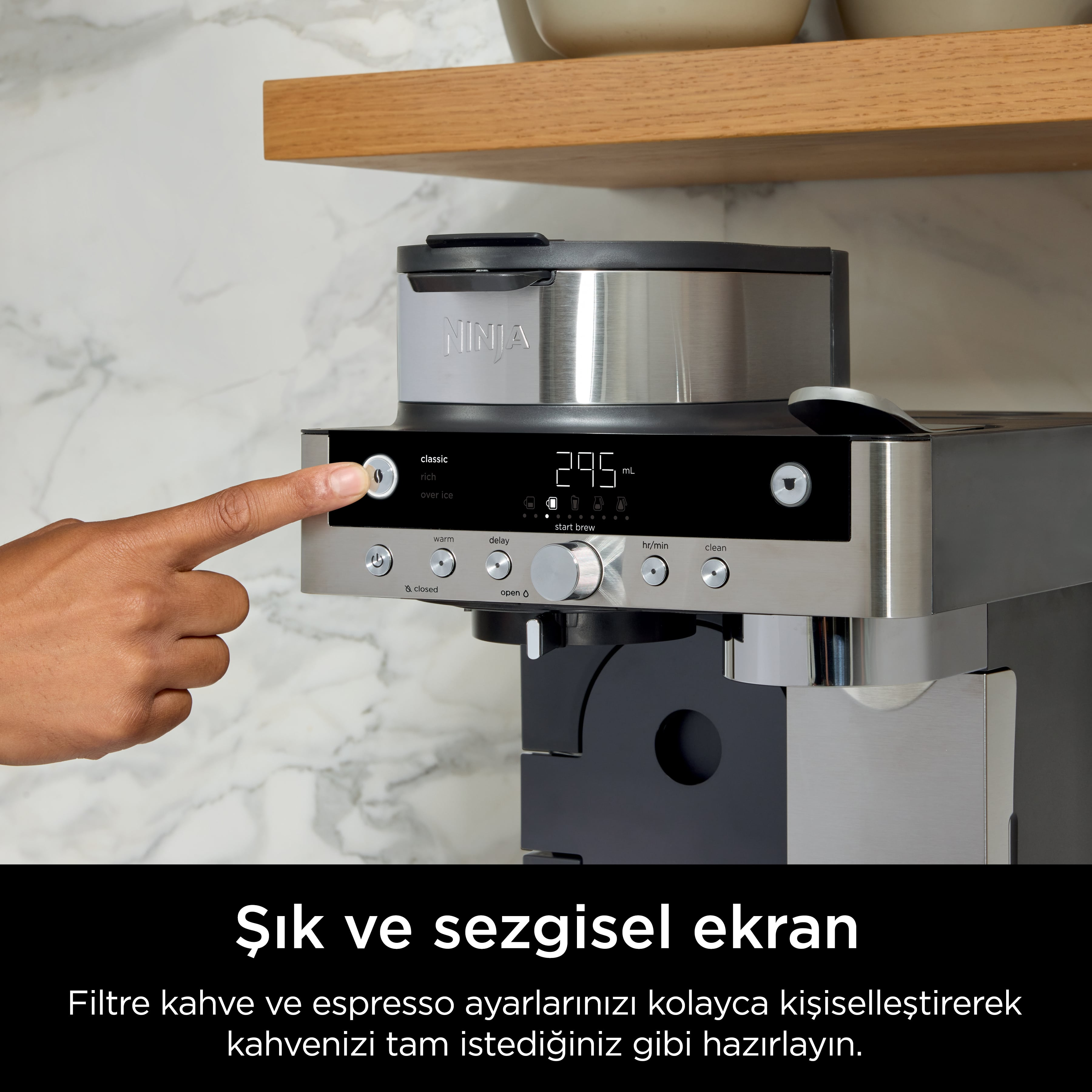 Bir el, kahve makinesinde bir düğmeye basıyor. Makine, ayarları gösteren siyah bir ekrana sahip gümüştür.