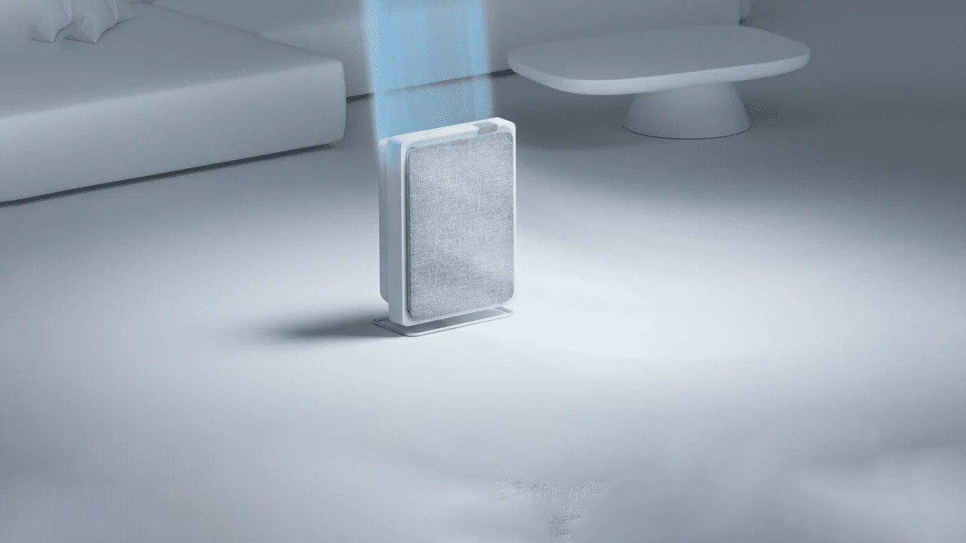 Oczyszczacz powietrza stoi w jasnym pokoju. Łóżko i stół są w tle. Niebieska wiązka światła świeci z oczyszczacza.