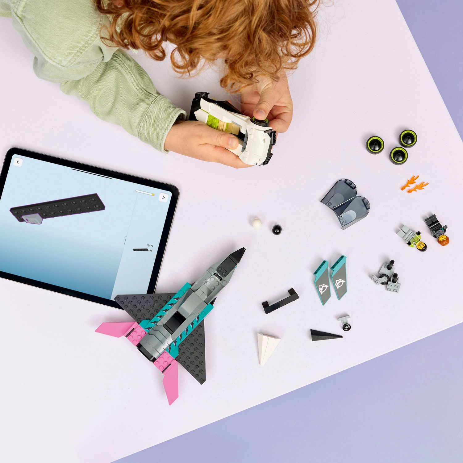 Un bambino sta costruendo un aereo e un'auto giocattolo. I pezzi e le istruzioni sono su una superficie bianca e viola, con un tablet nelle vicinanze.