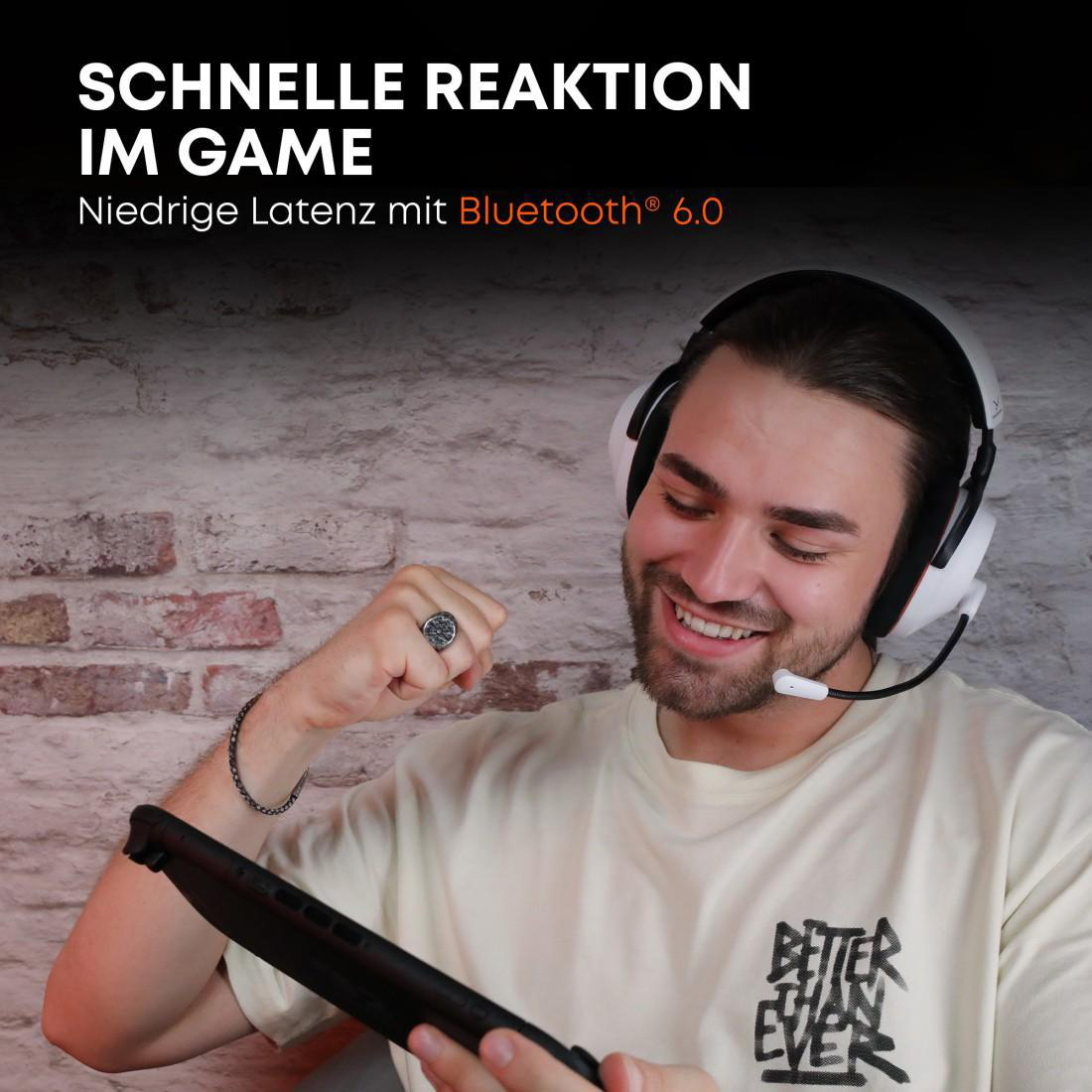 Ein Mann mit Headset und Mikrofon hält eine Handheld-Konsole und lächelt. Im Hintergrund eine Backsteinmauer.