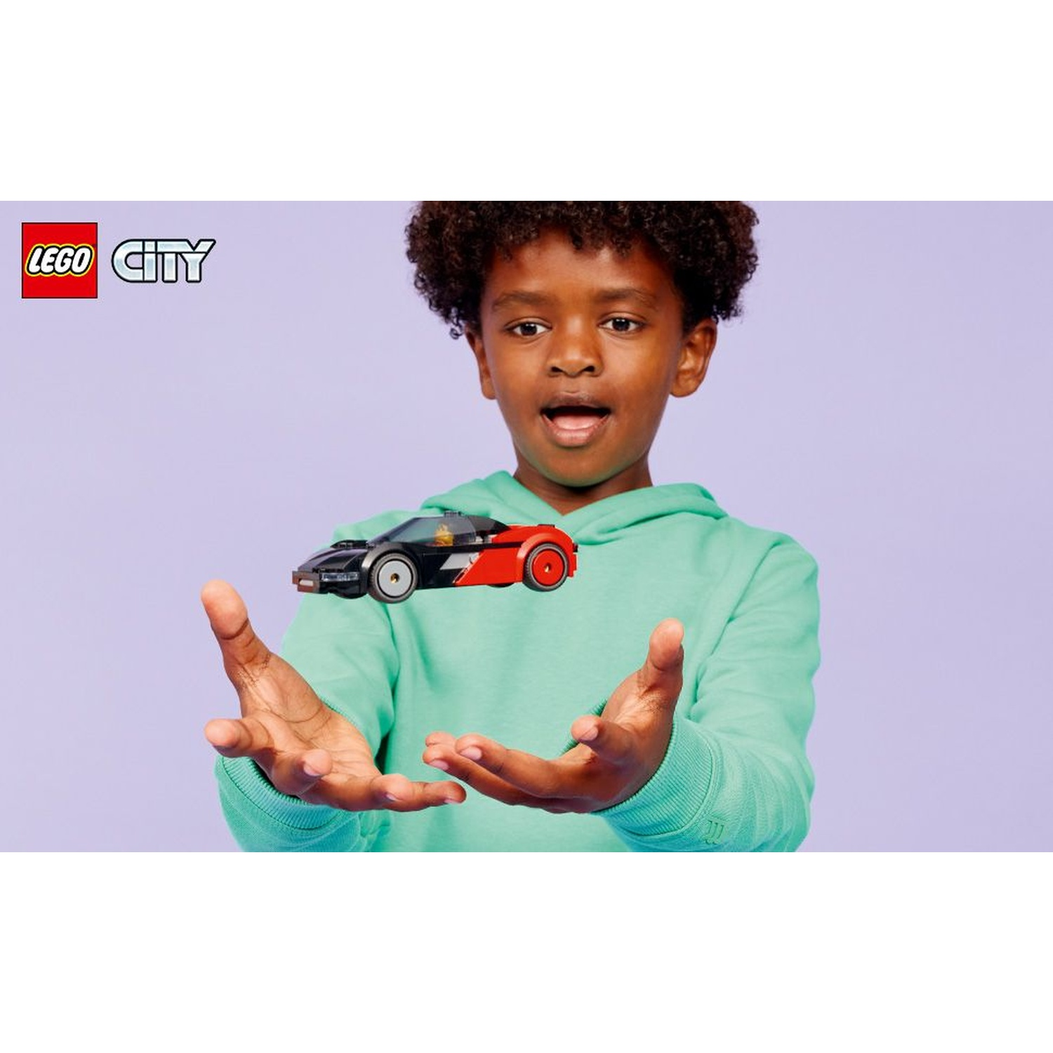 Un ragazzino con una felpa verde con cappuccio tiene in mano un'auto LEGO. L'auto è nera e rossa. Lo sfondo è lilla chiaro.
