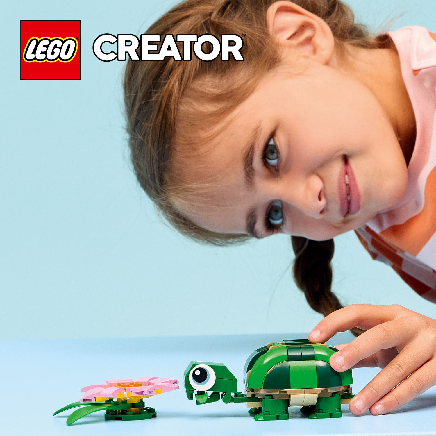 Una ragazza guarda una tartaruga e un fiore Lego. La tartaruga è verde, il fiore rosa e giallo. Lo sfondo è azzurro.