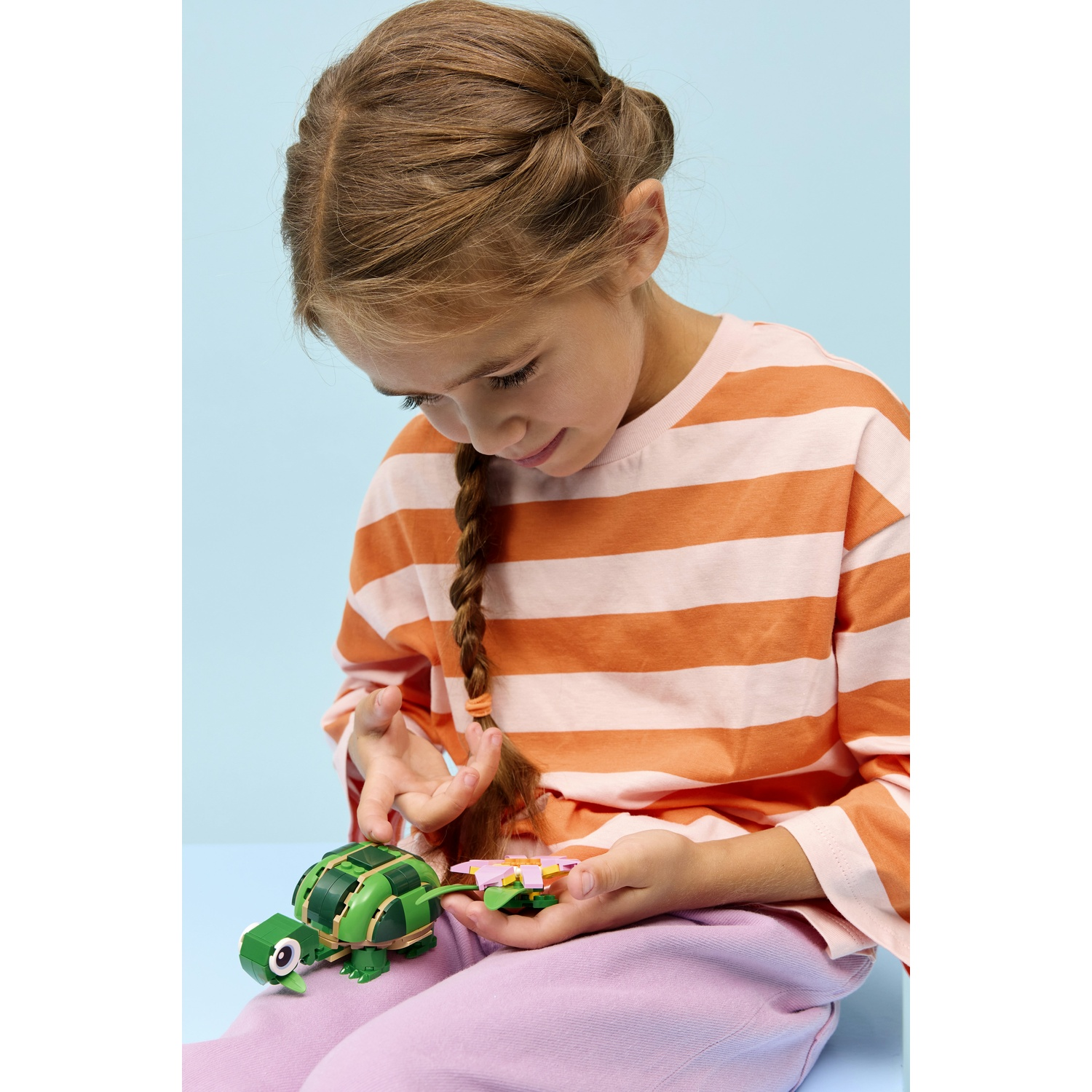 Una ragazza con una treccia tiene in mano una tartaruga giocattolo verde e un fiore rosa su sfondo blu, con una maglia a righe arancioni e bianche.