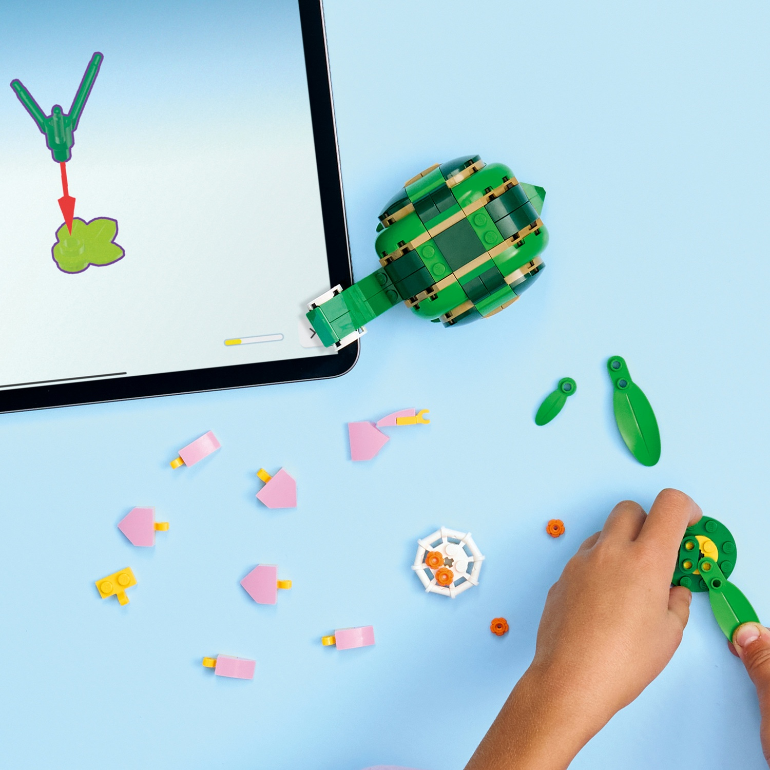 Una persona assembla un carciofo Lego verde su una superficie blu, con pezzi sparsi e un tablet che mostra le istruzioni.